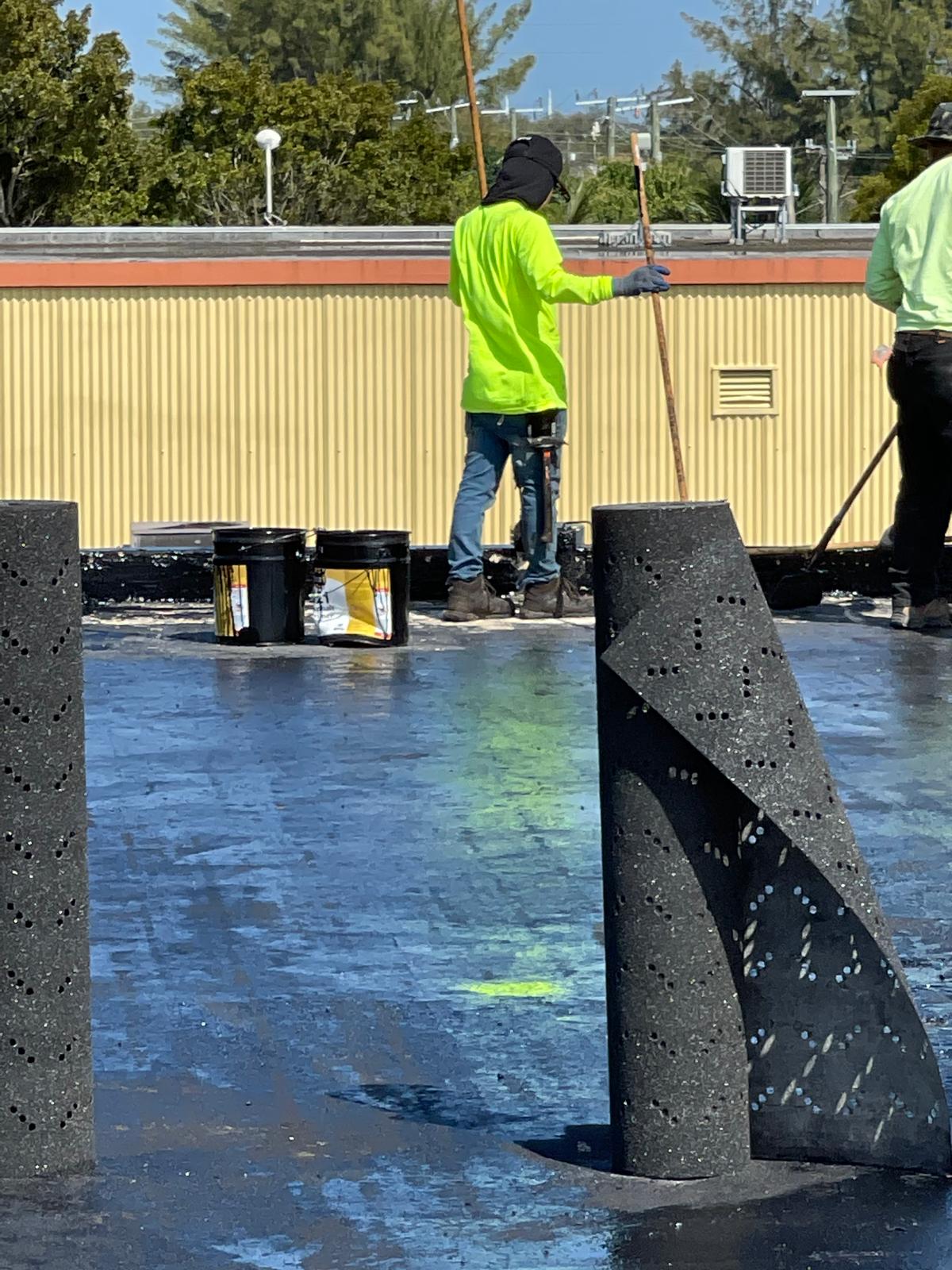 Two workers in neon shirts apply roofing material on a flat roof under a sunny sky, with tools and buckets nearby.