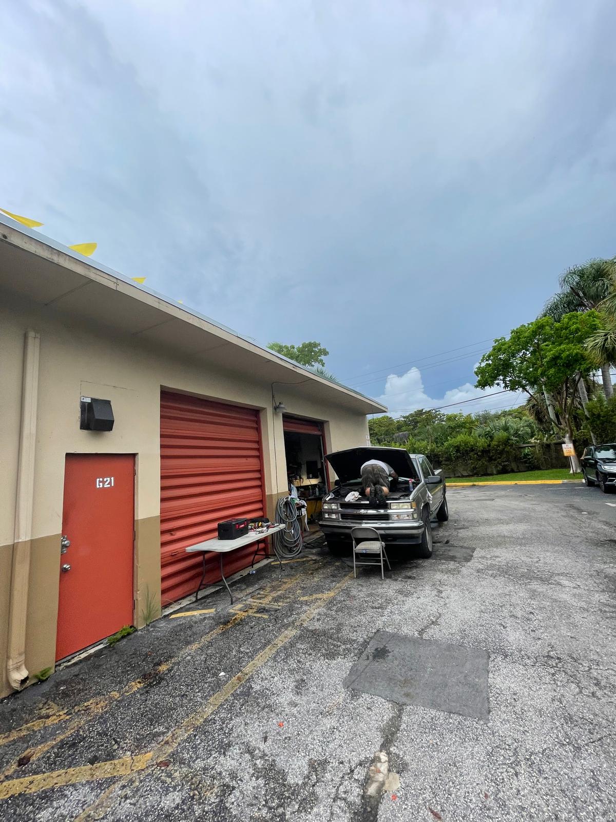 Person working under the hood of a car outside a garage with a red door and rolling shutter.