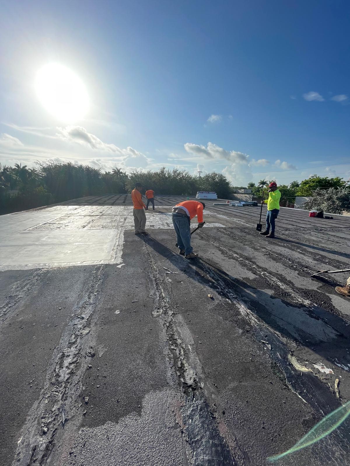 Four workers in bright shirts repair a flat rooftop under a clear, sunny sky with trees in the background.