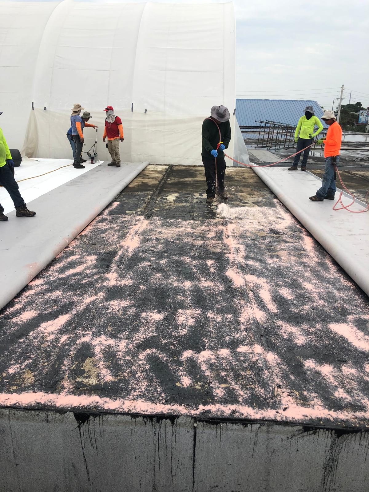 Workers apply a pink substance on a flat roof while others stand nearby, with white tarp structures in the back.