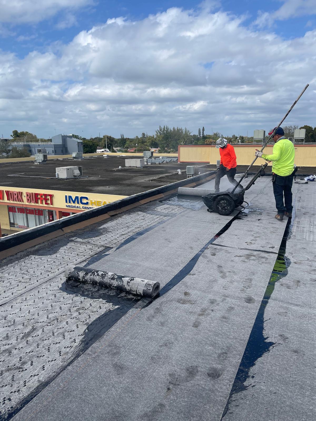 Two workers apply roofing material on a flat roof under a partly cloudy sky, with buildings in the background.