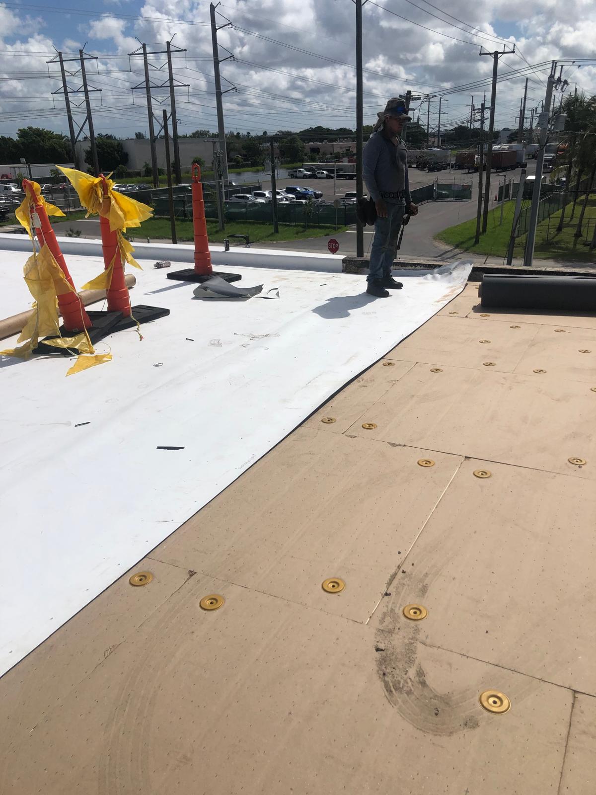 A worker installing a white membrane roof, with cones and traffic in the background on a sunny day.