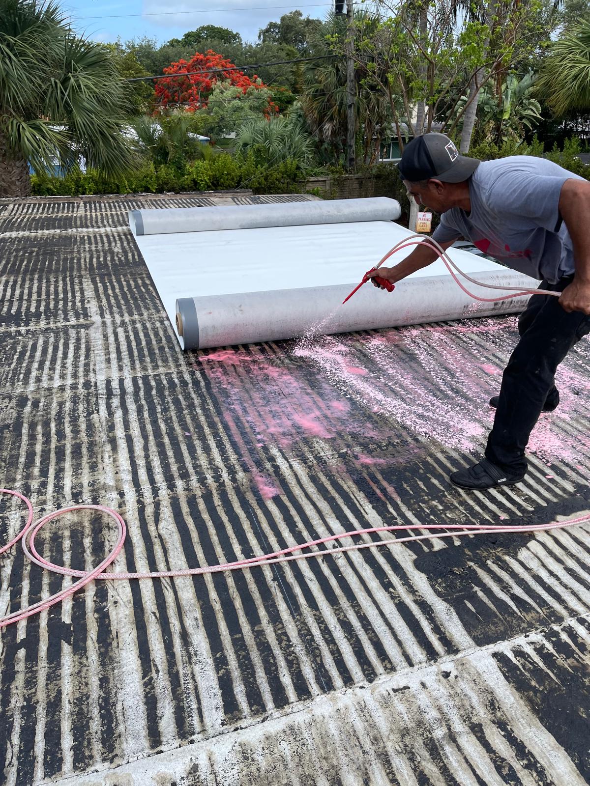 A worker sprays pink adhesive on a striped rooftop, preparing to roll out roofing material.