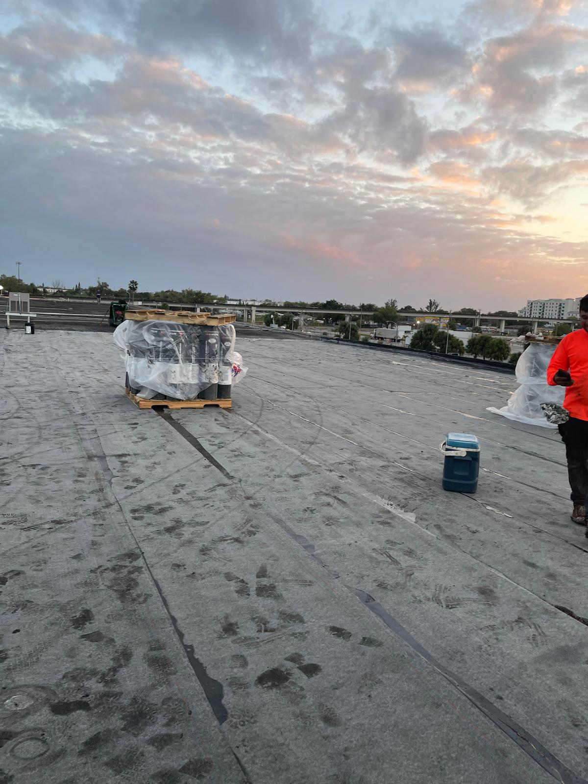 A rooftop at sunset with pallets of materials, a person in an orange shirt, and a cloudy sky.