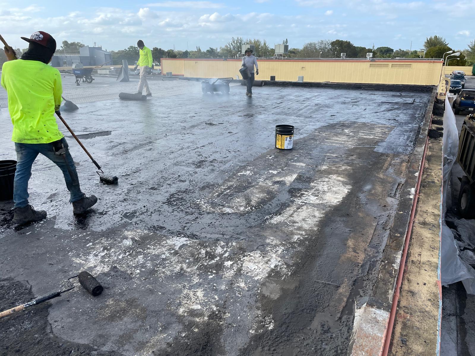 Workers in safety gear repair and seal a flat commercial roof with tools and buckets on a sunny day.