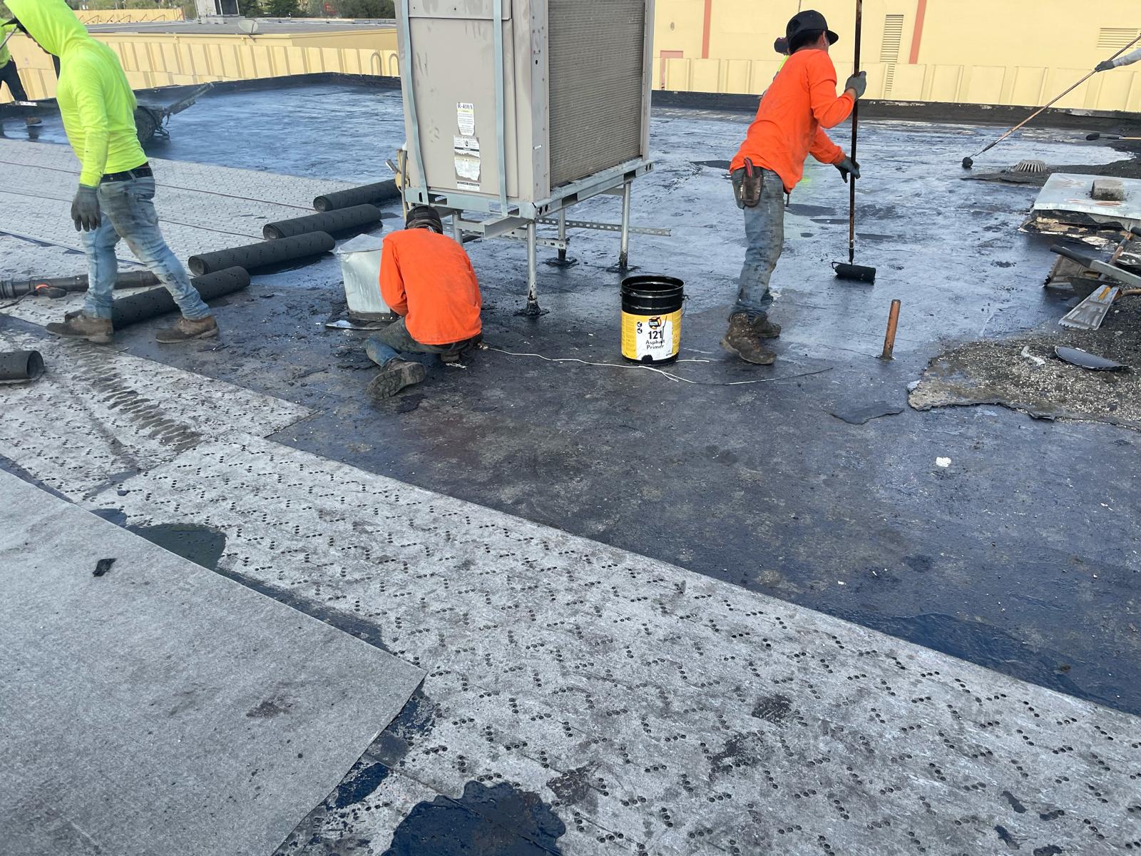 Three workers repairing a flat rooftop with tools, black sealant, and roofing materials near an HVAC unit.