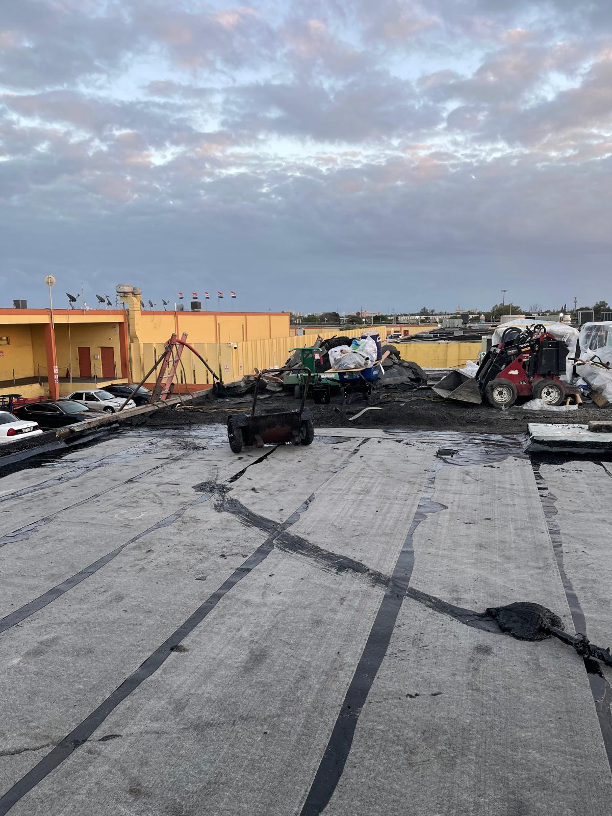 Damaged rooftop with debris, machinery, and construction materials under a cloudy sky near yellow buildings.