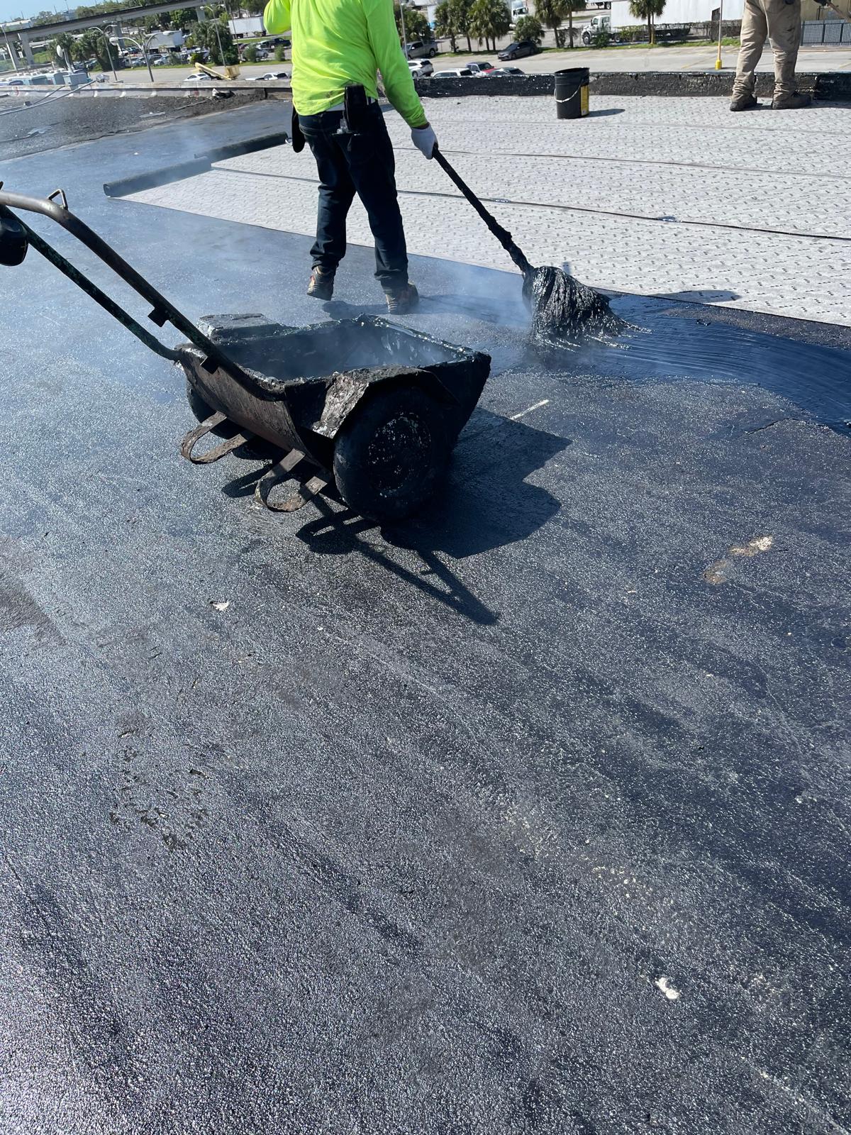 Worker in a neon shirt spreads hot tar on a flat roof using a mop; tar cart and steam visible.