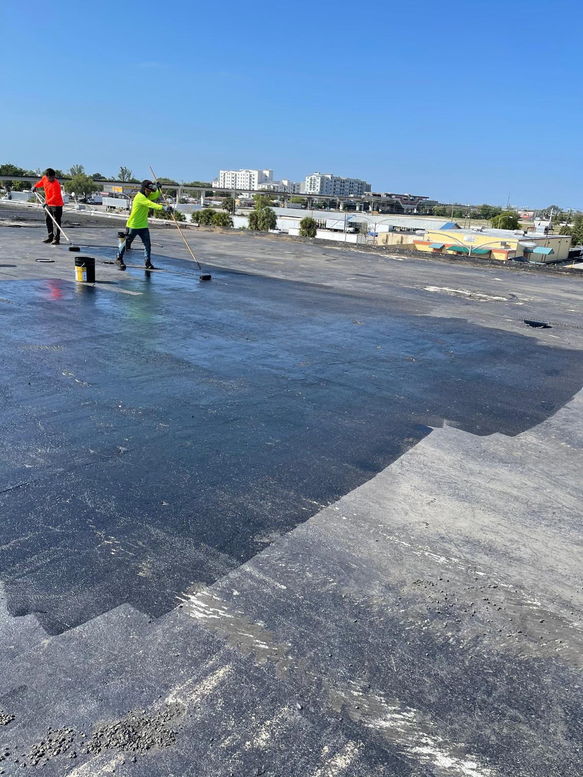 Two workers in bright shirts apply coating to a large, flat rooftop under a clear blue sky.