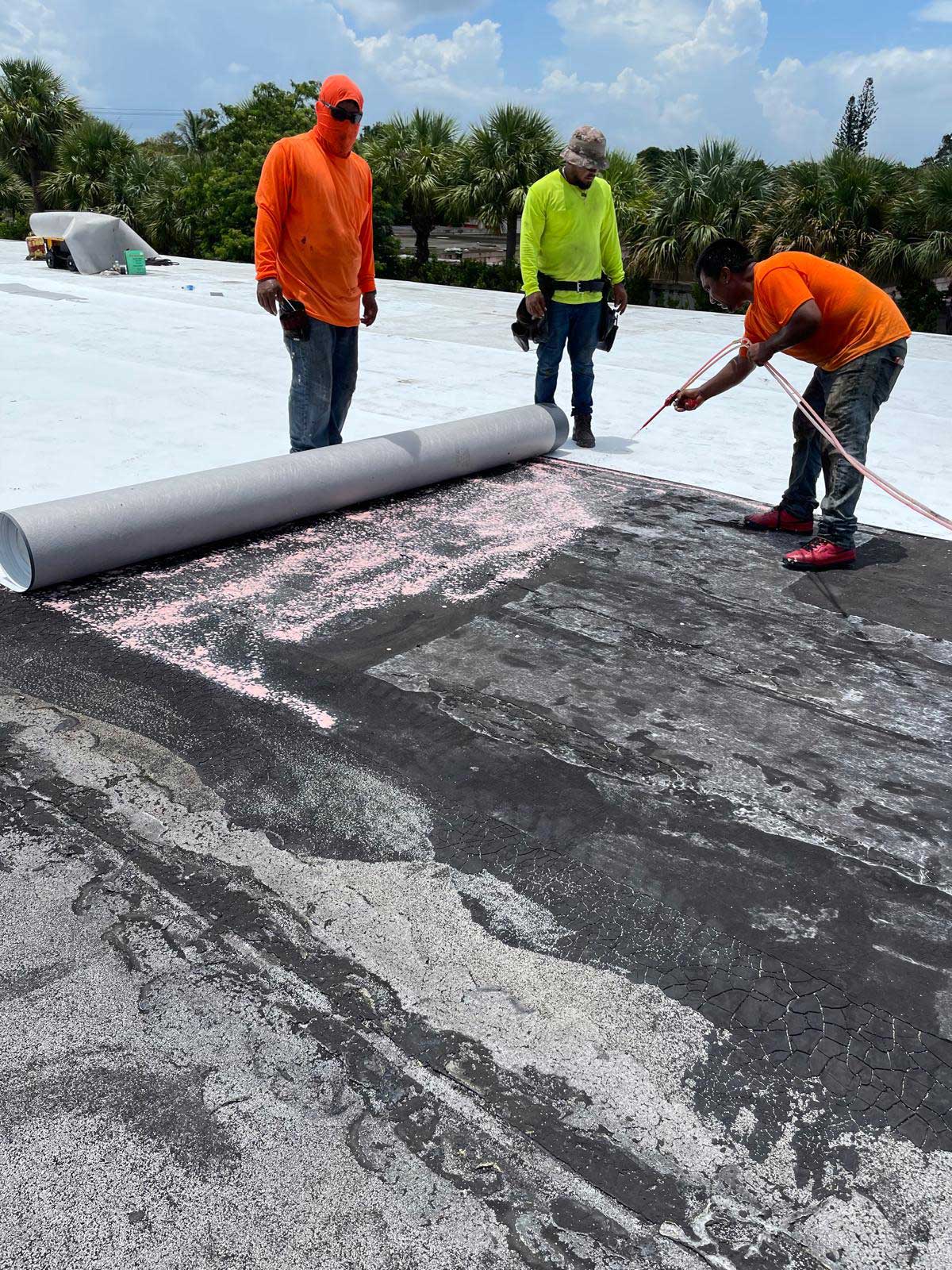 Three workers in bright shirts roll out roofing material on a flat roof under a sunny sky with palm trees nearby.