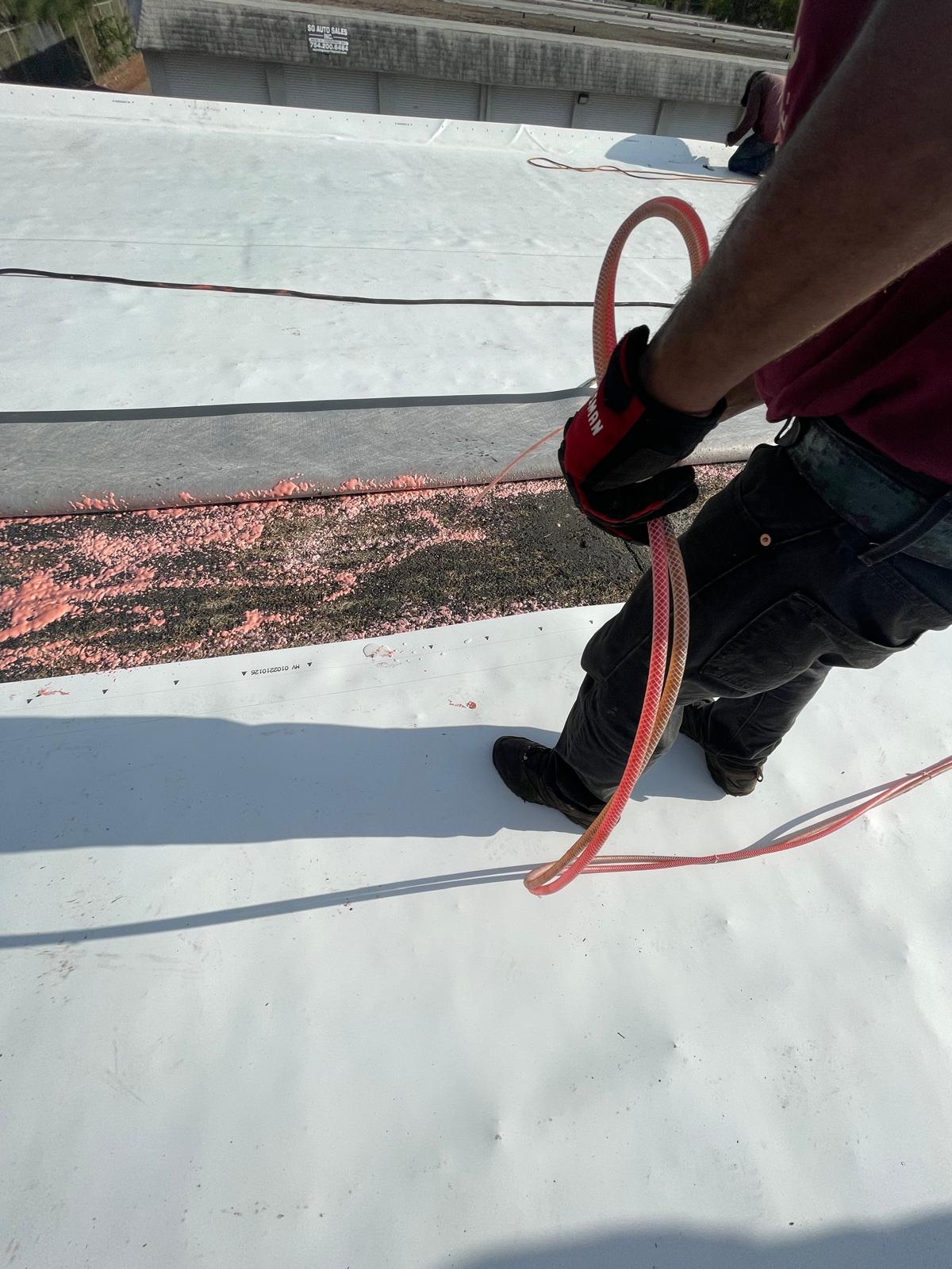 Person in gloves holding a hose, standing on a white roof membrane near scattered pink foam or insulation.