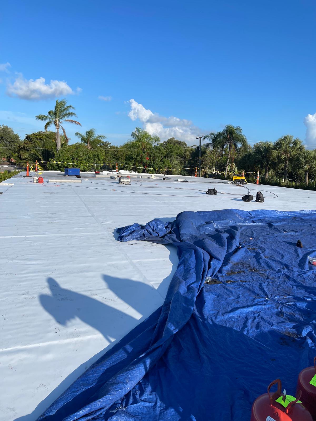 A rooftop under construction with a blue tarp, palm trees, and construction materials under a clear sky.