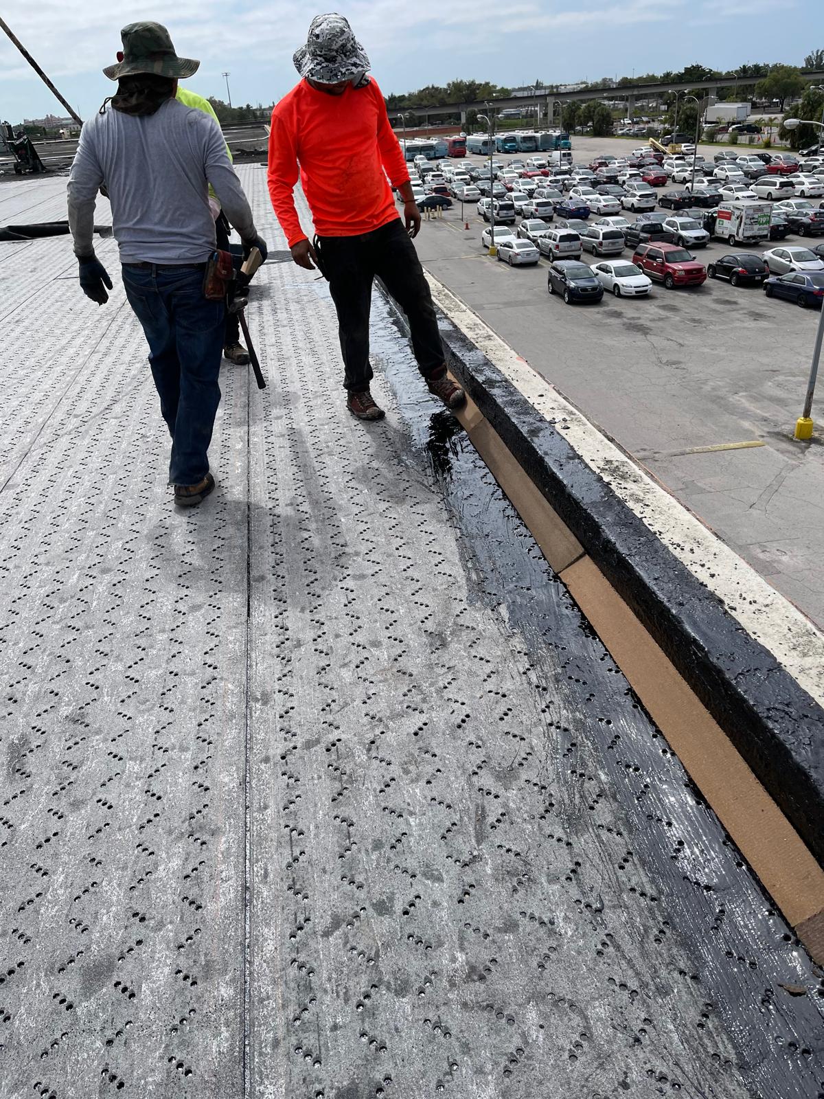Two workers on a rooftop near the edge, overlooking a parking lot filled with cars on a cloudy day.
