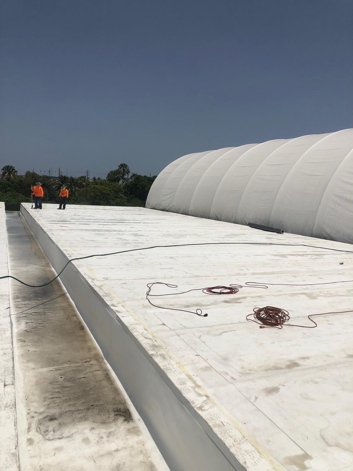 Two workers in orange shirts stand on a large white rooftop near a dome-shaped structure under a clear sky.