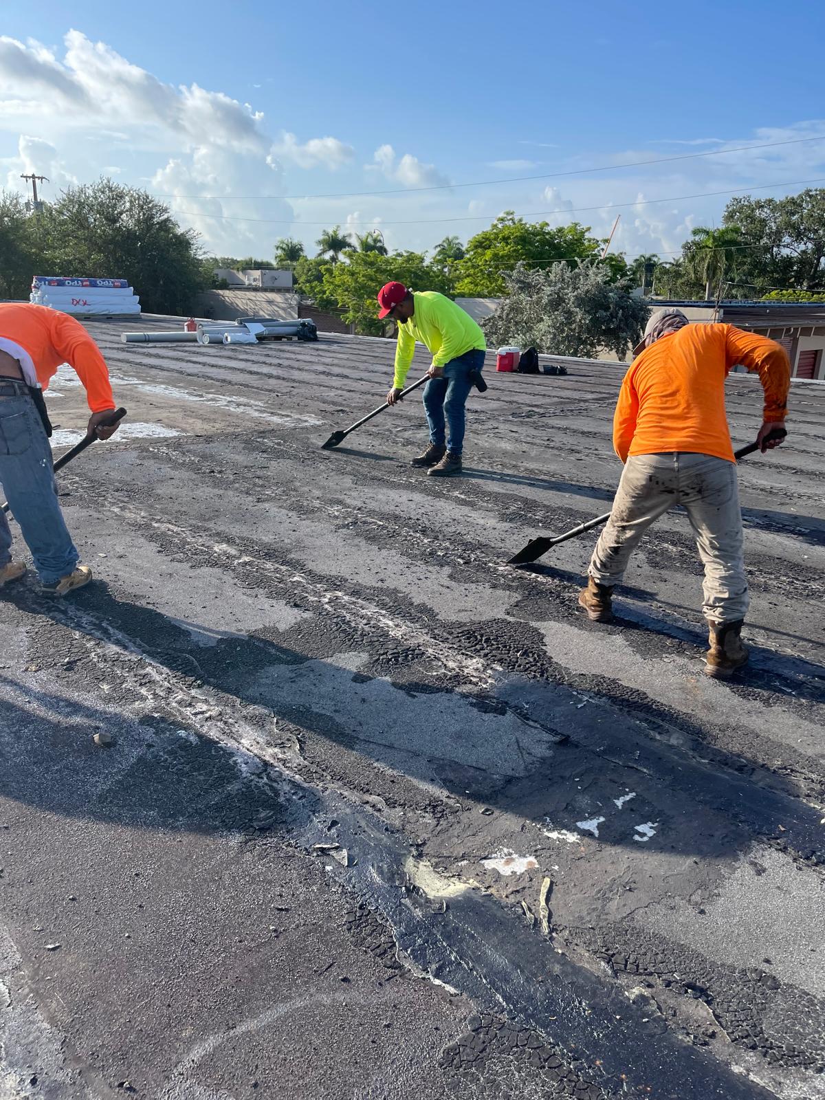 Three workers in bright shirts clear debris from a flat rooftop under a sunny, blue sky.