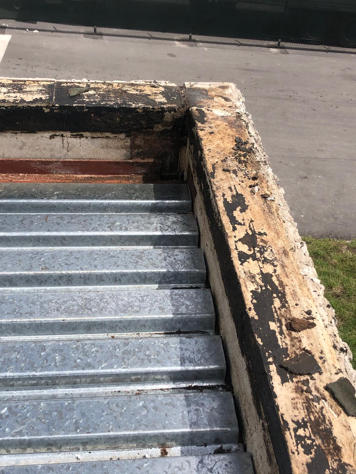 Corrugated metal roof edge with weathered, peeling paint and cracked concrete, next to pavement and grass.