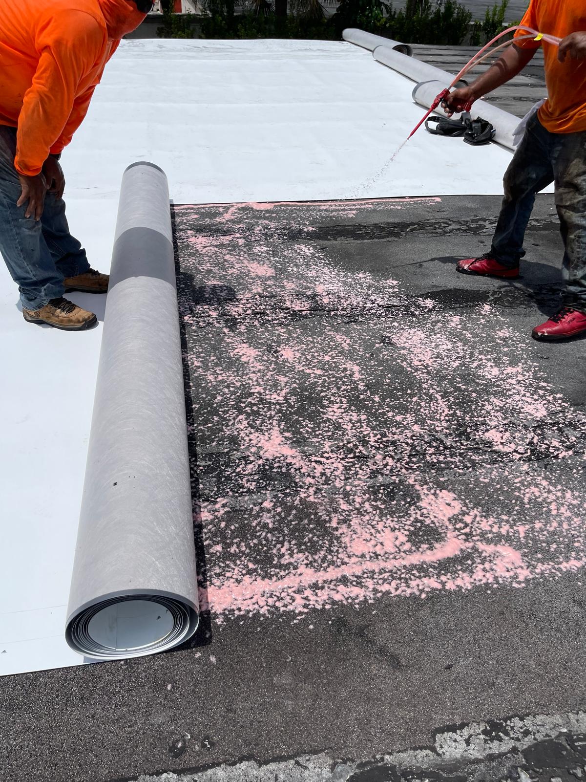 Two workers unrolling roofing material over adhesive on a flat roof, with pink spray visible on the surface.