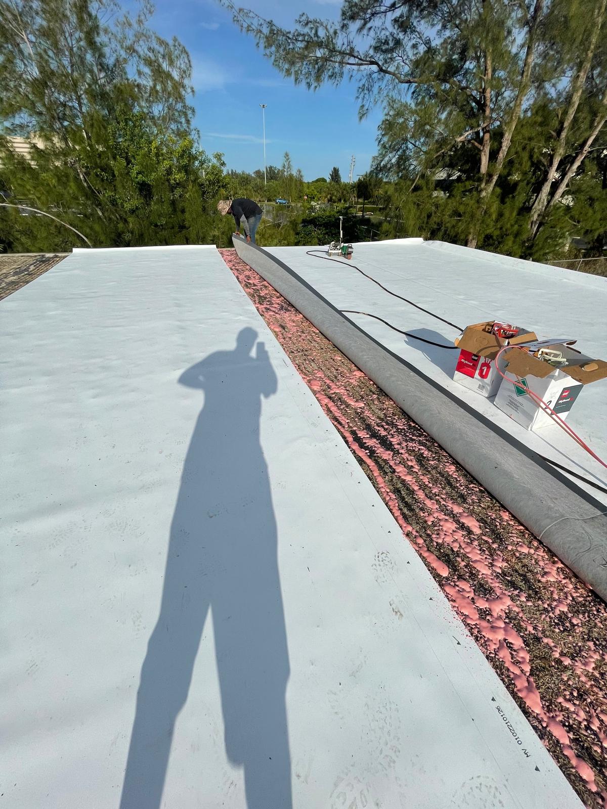 Person working on a flat rooftop with roofing materials; long shadow cast in the foreground.