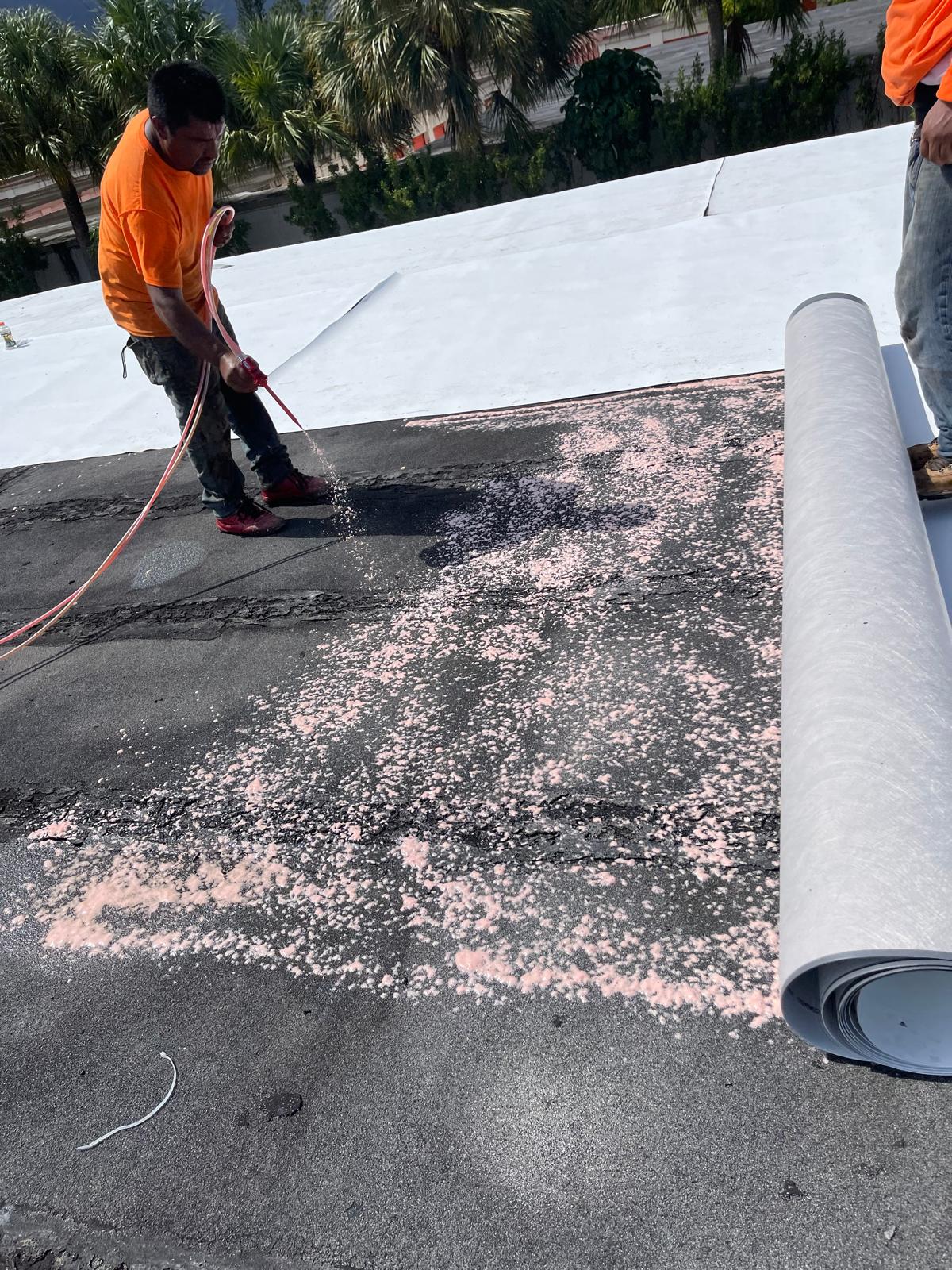 Worker in orange shirt spraying adhesive on a flat roof with roofing material unrolled beside him.