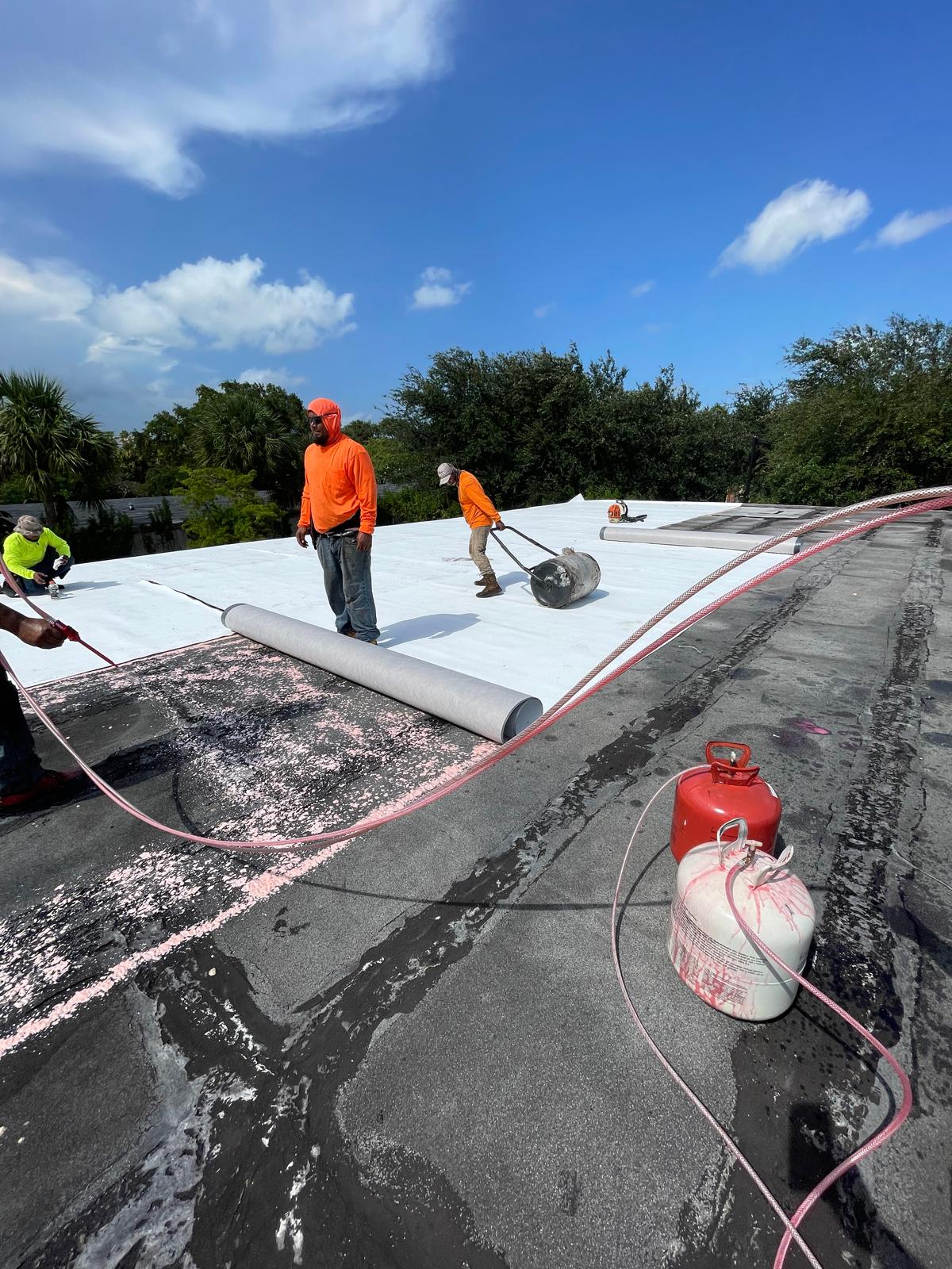 Three workers in orange shirts install white roofing material on a flat roof under a blue sky.