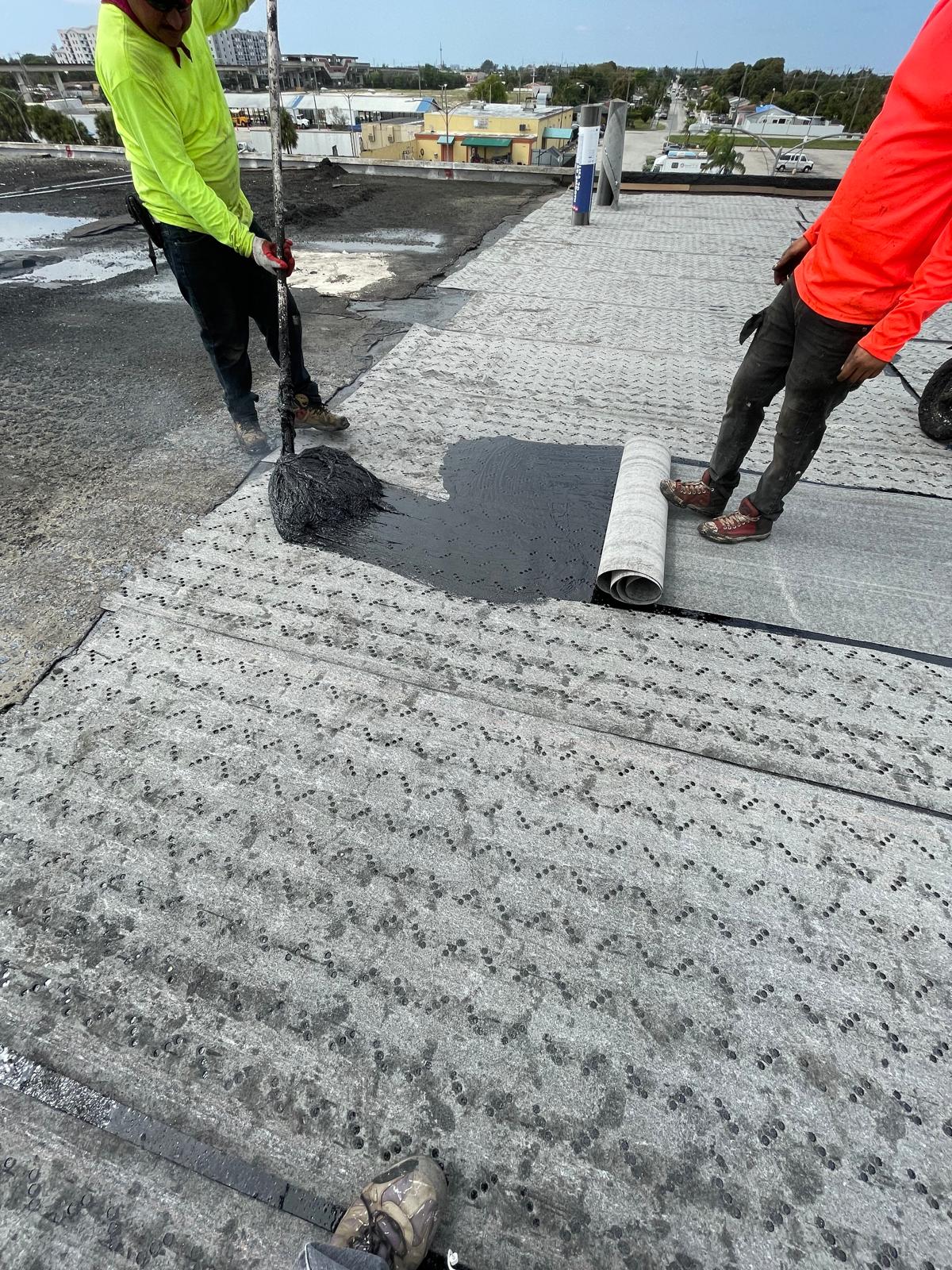 Two workers apply black adhesive to a rooftop while rolling out roofing material.