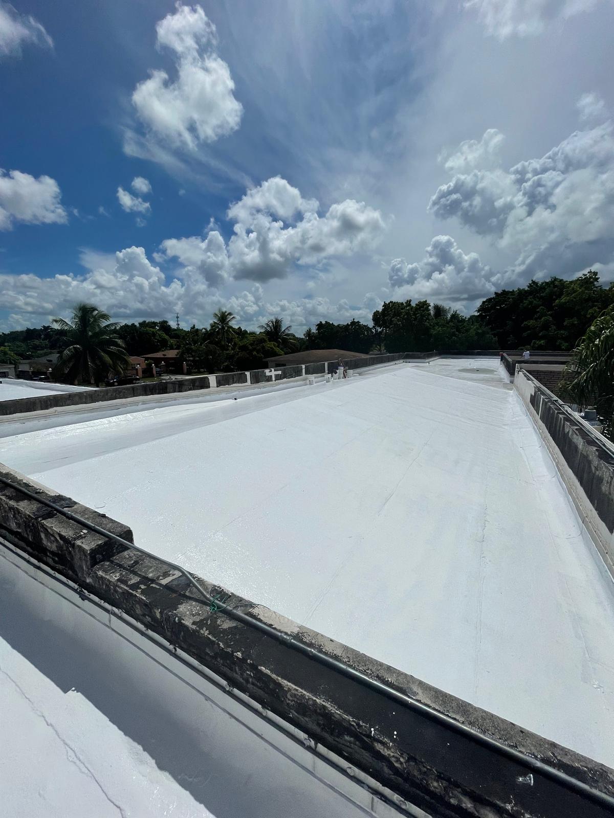 A white flat rooftop under a bright blue sky with scattered clouds and trees in the background.