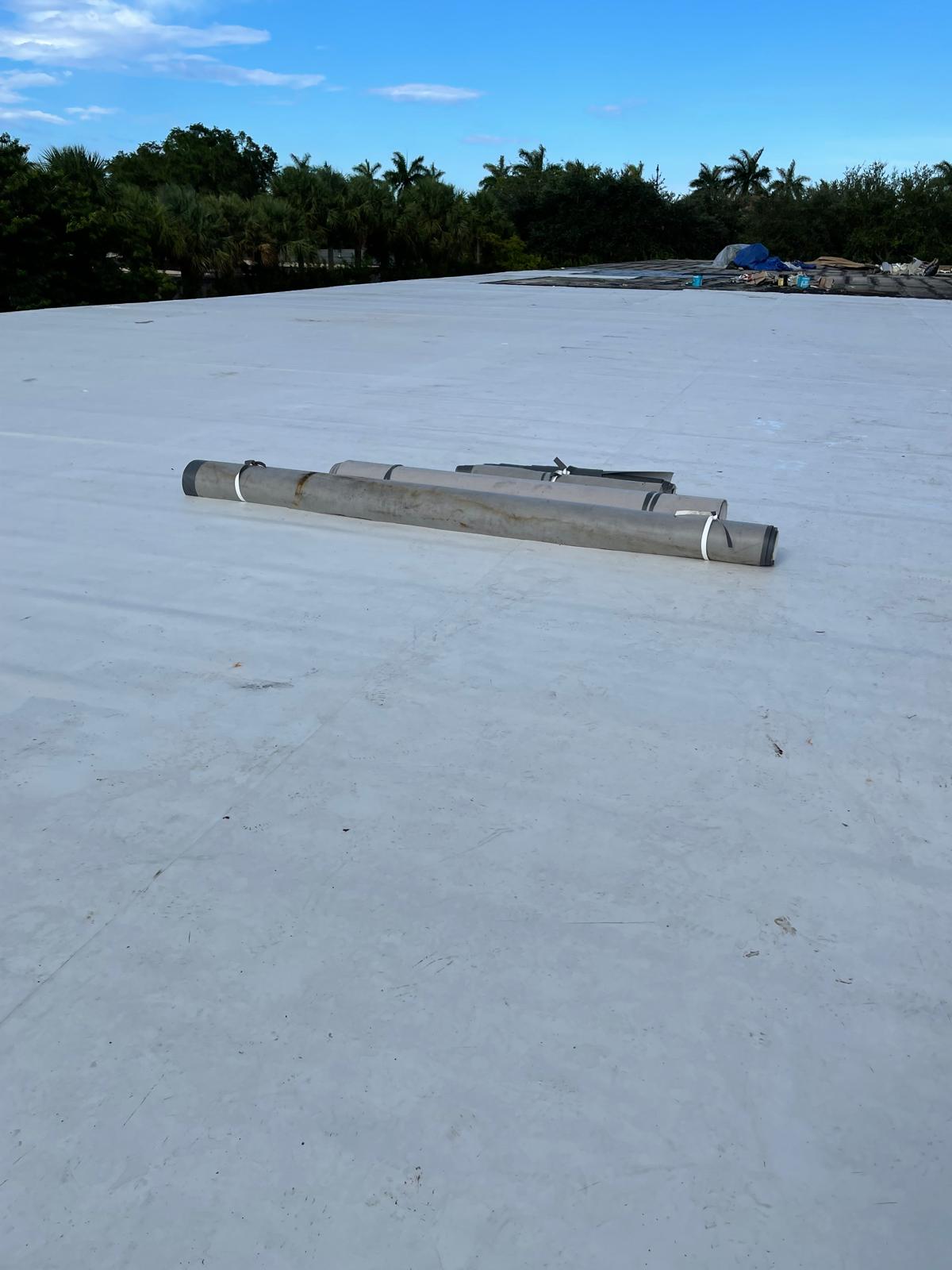 Large white rooftop with three rolled materials in the center; trees and blue sky in the background.
