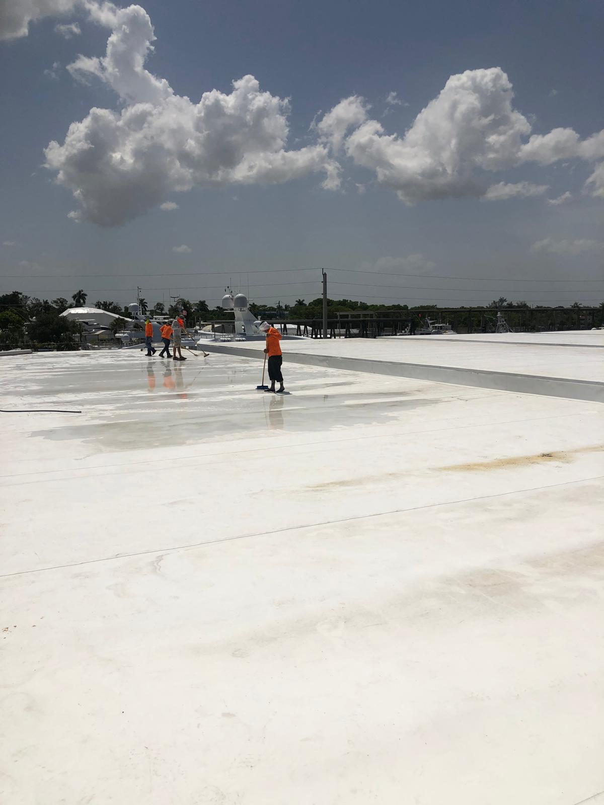 Workers in orange shirts clean a large white rooftop under a partly cloudy sky.