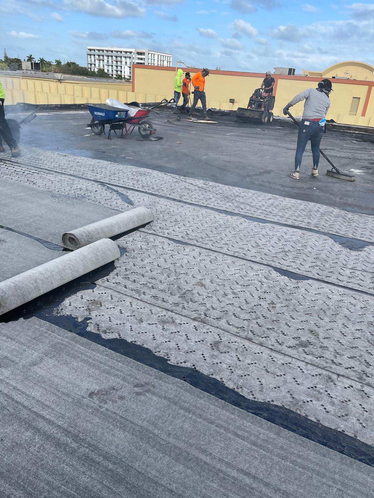 Workers installing roofing material on a flat rooftop under a partly cloudy sky; tools and equipment visible.