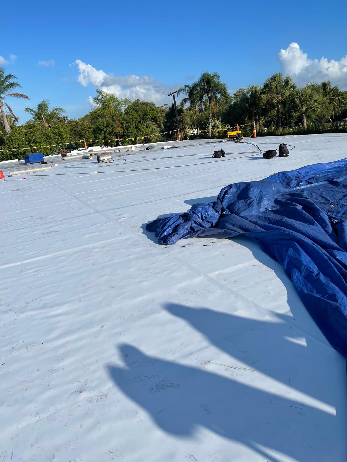 Large white rooftop under construction with tools and a blue tarp, surrounded by palm trees in sunlight.