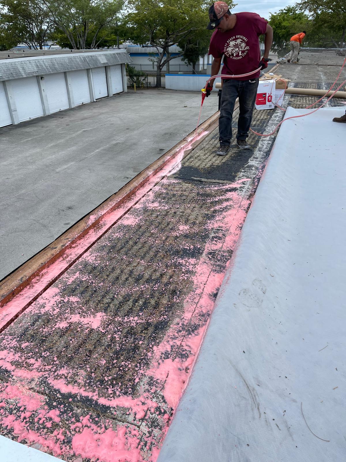 Worker in maroon t-shirt sprays sealant on a flat rooftop with exposed pink insulation and gravel.