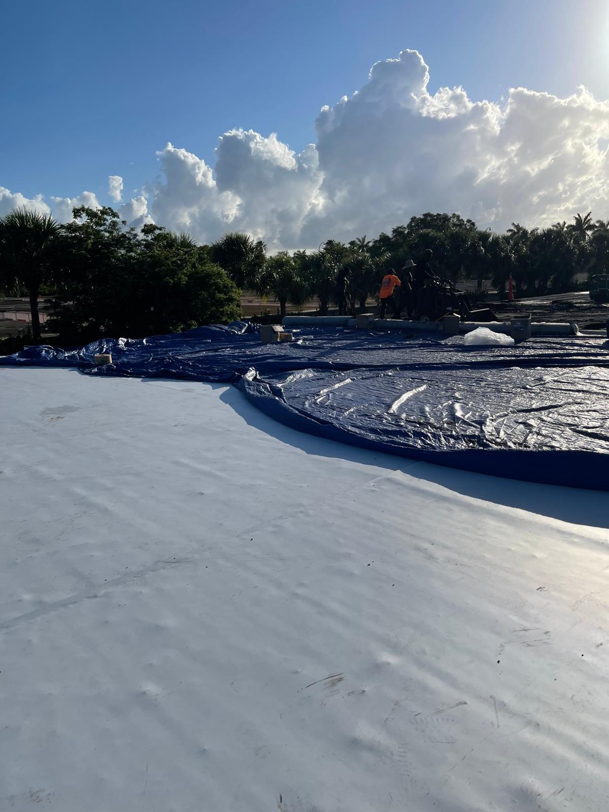 A worker stands near a large blue tarp covering part of a construction site under a bright, cloudy sky.