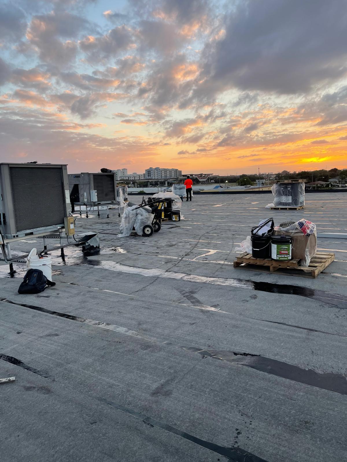 A rooftop with construction materials and equipment at sunset, with a person in an orange vest in the distance.