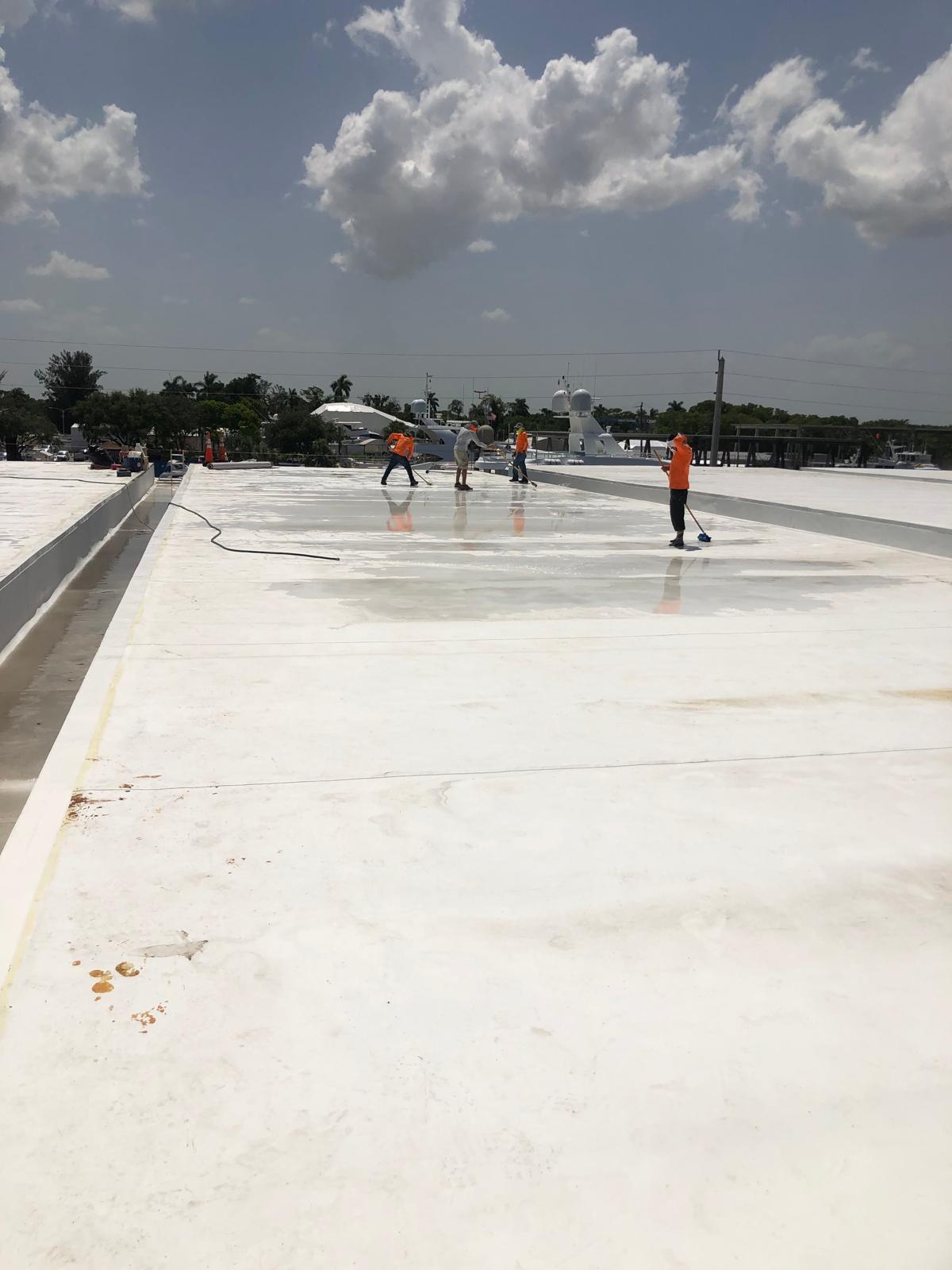 Four workers in orange shirts clean or coat a large flat white rooftop under a partly cloudy sky.