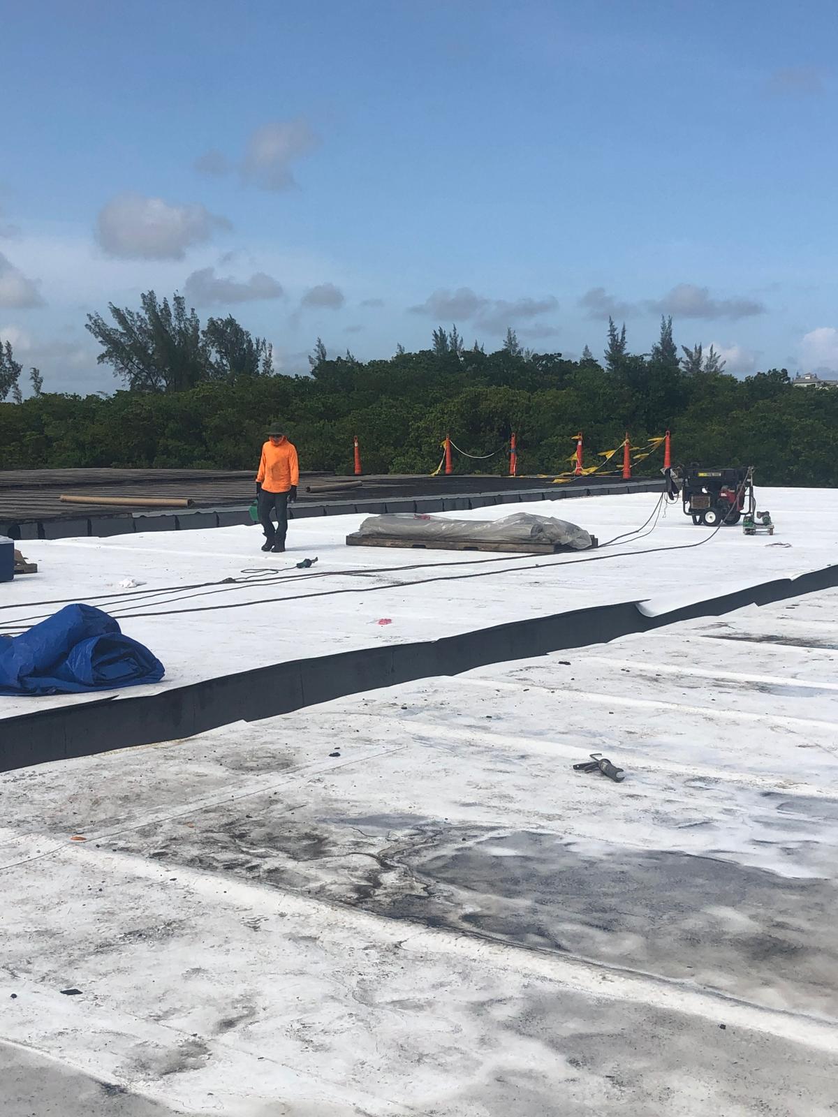 Person in an orange shirt works on a flat white rooftop with tools and equipment, trees in the background.