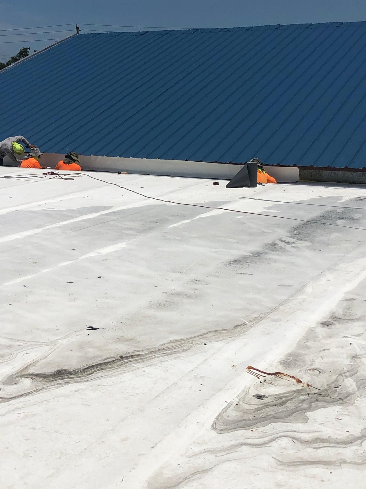 Workers in safety gear install roofing material on a white roof next to a sloped blue metal roof.