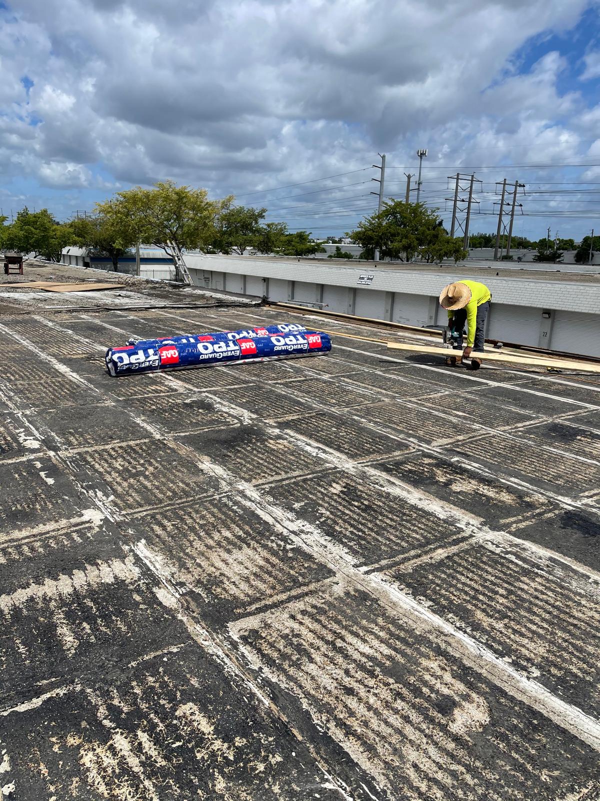 A worker in a hat prepares materials on a flat, weathered rooftop under a partly cloudy sky.