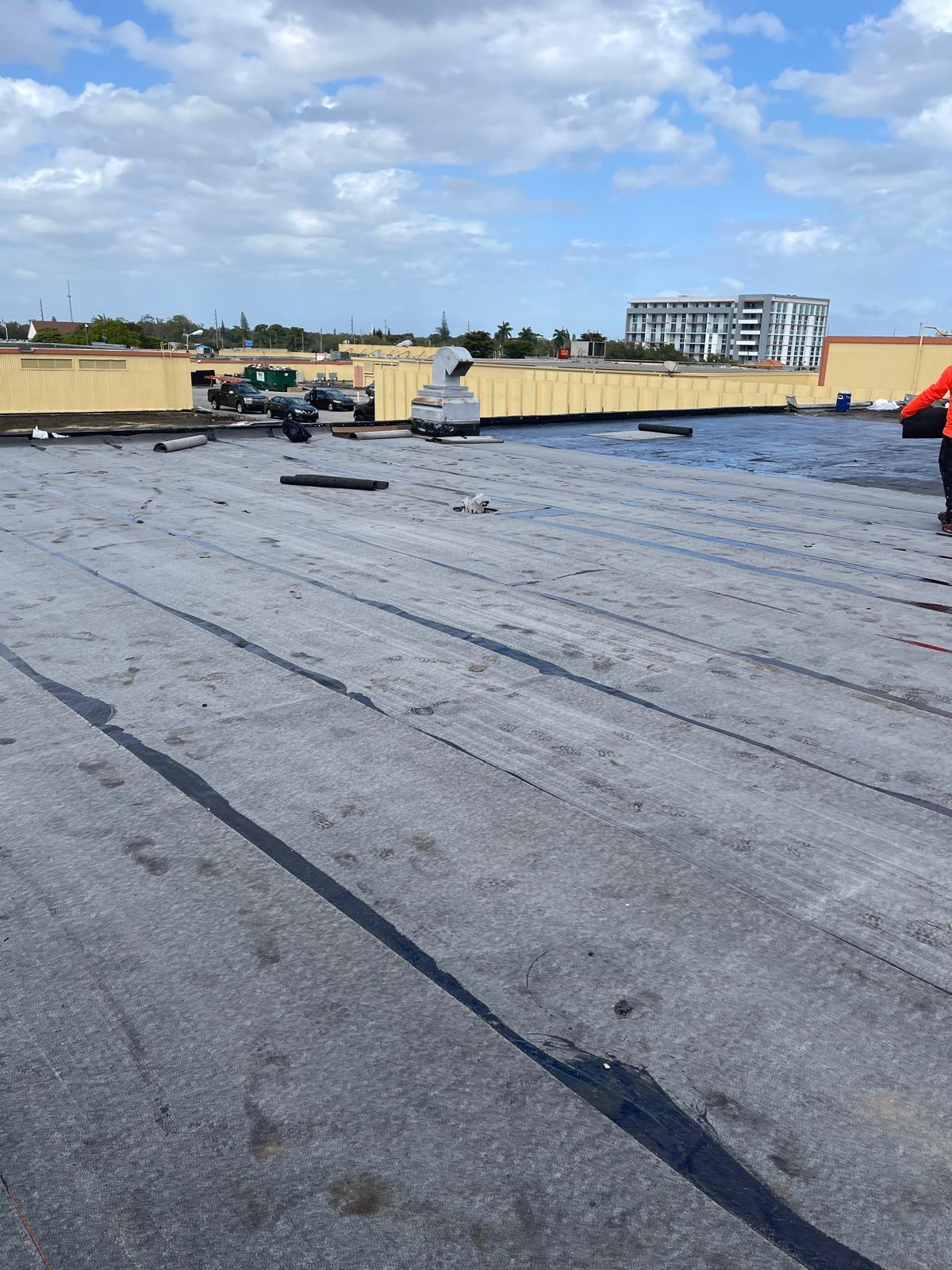 A large flat rooftop with black waterproofing material, some equipment, and cloudy blue sky overhead.