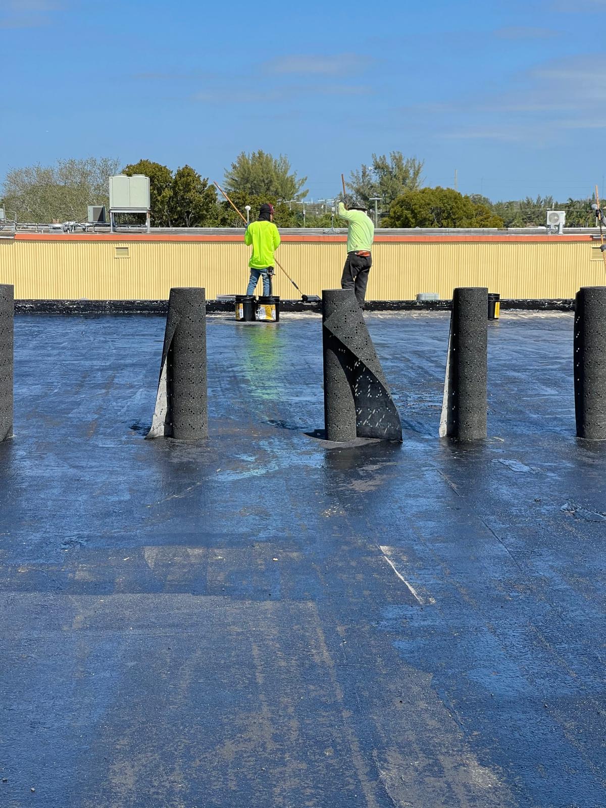 Workers in neon shirts apply roofing material on a flat roof under a clear blue sky.