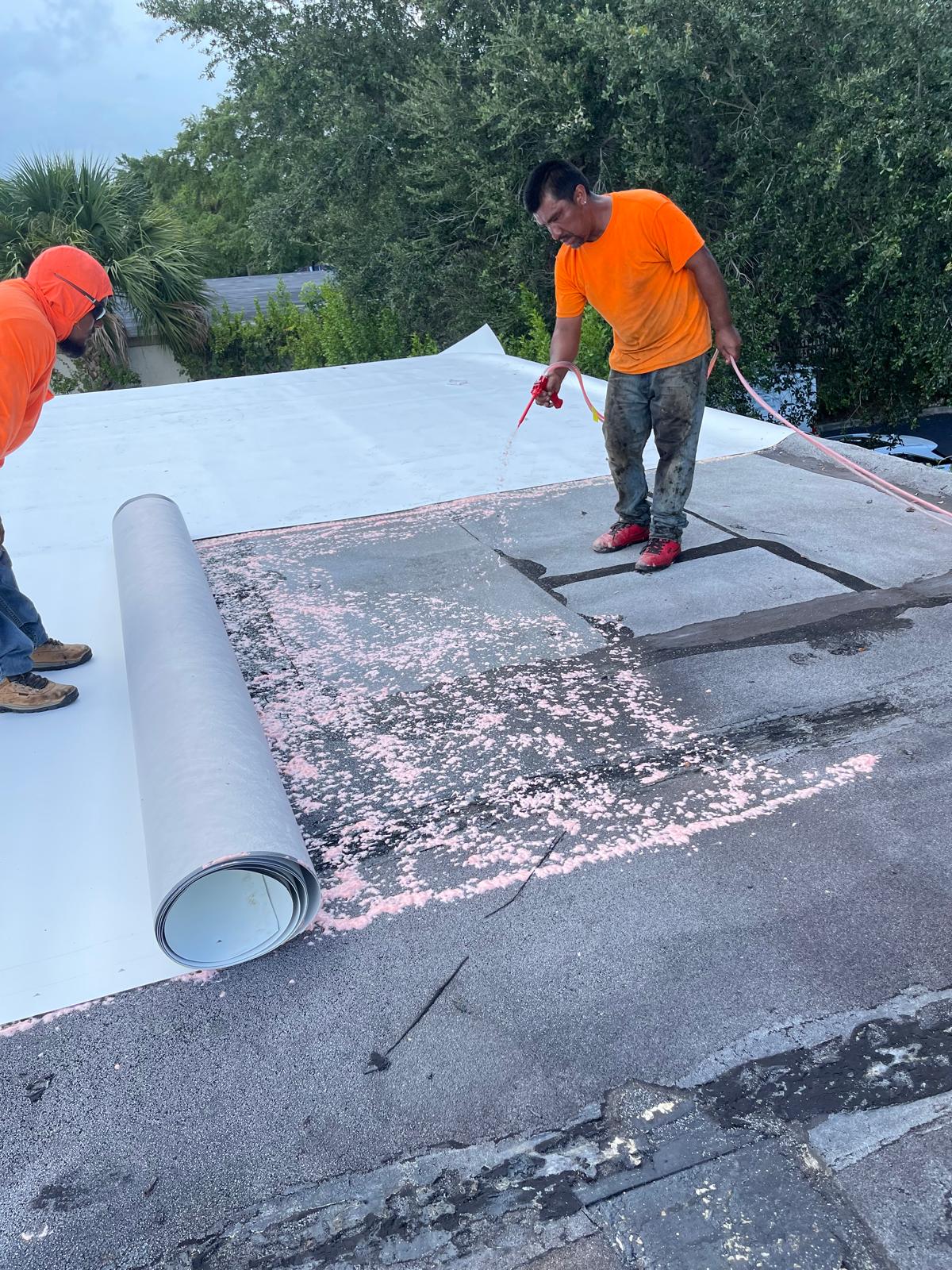 Two workers in orange shirts install roofing material, unrolling a sheet and spraying pink adhesive on the roof.