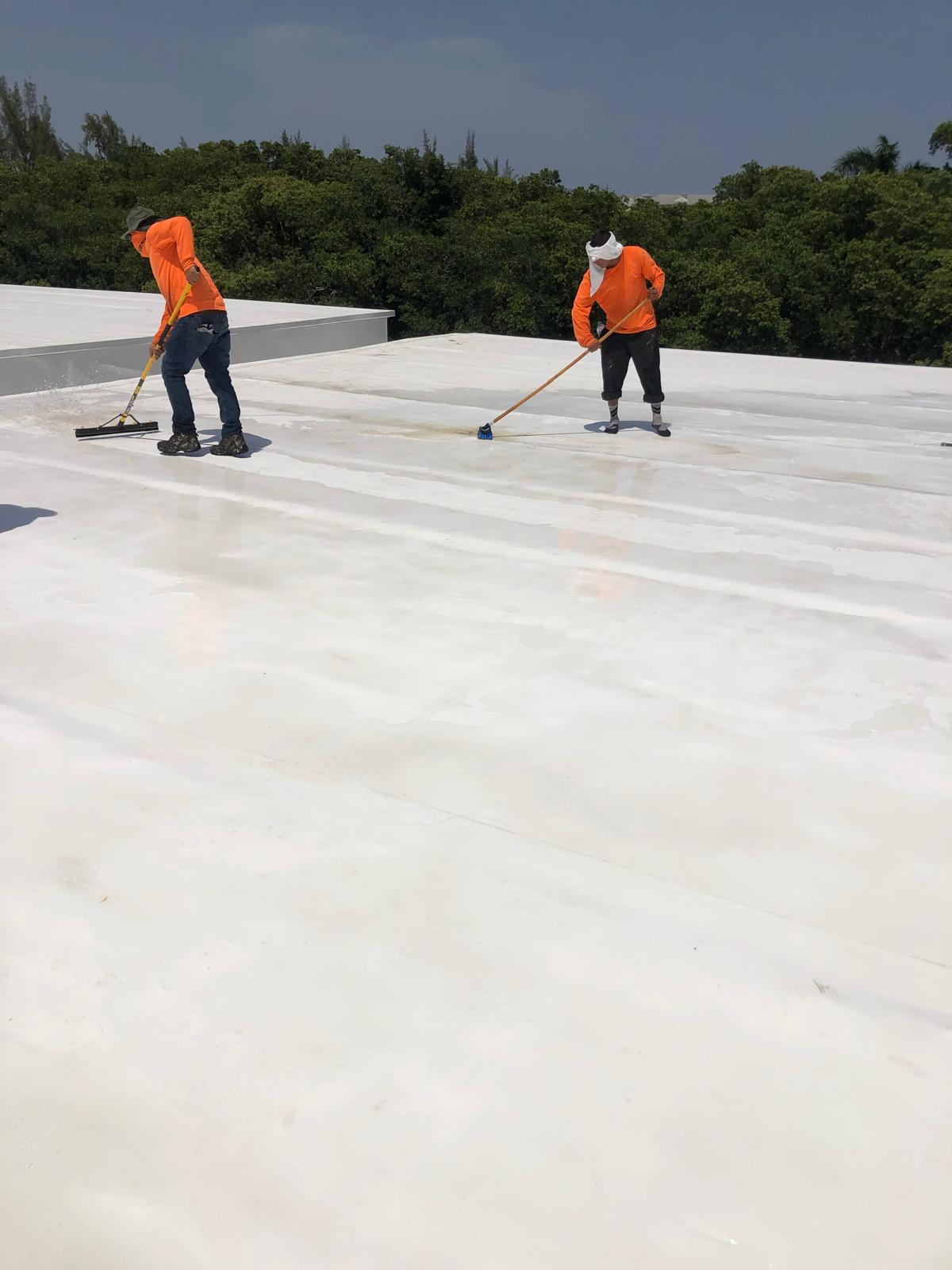 Two workers in orange shirts and hats apply coating to a flat white rooftop on a sunny day.