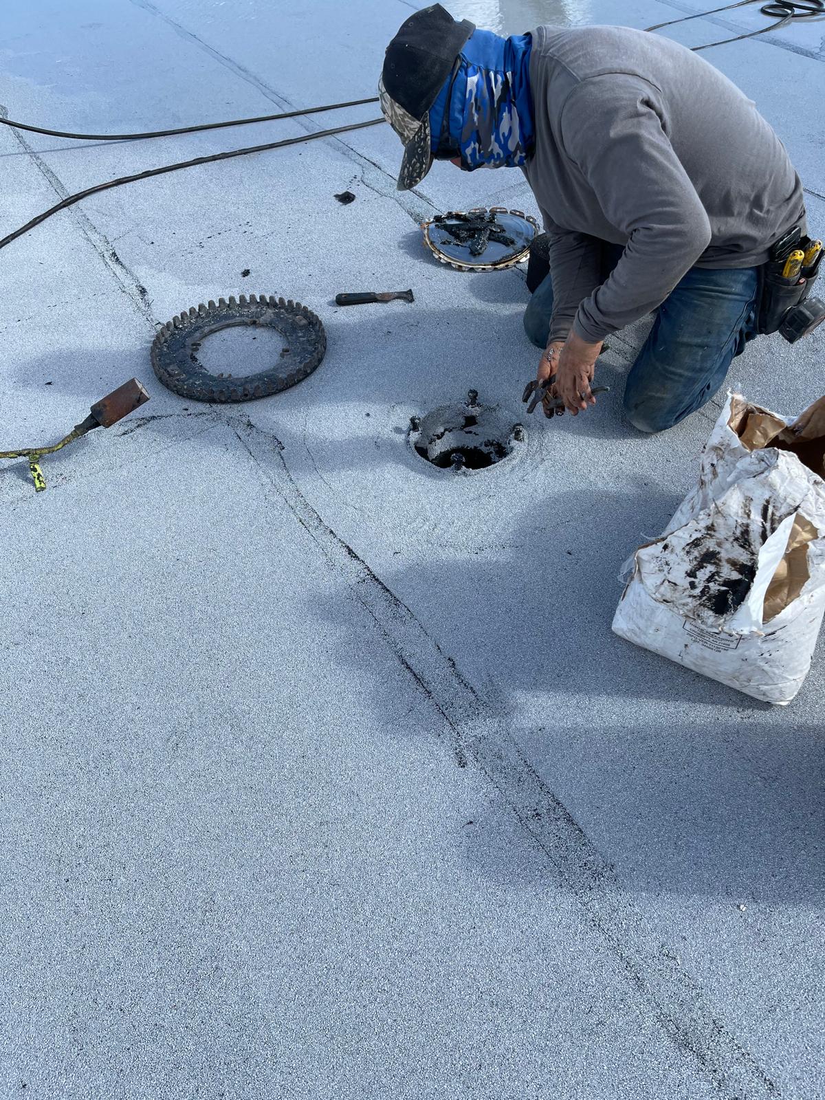 A worker kneels on a flat roof, repairing a drain with tools and a bag of materials nearby.