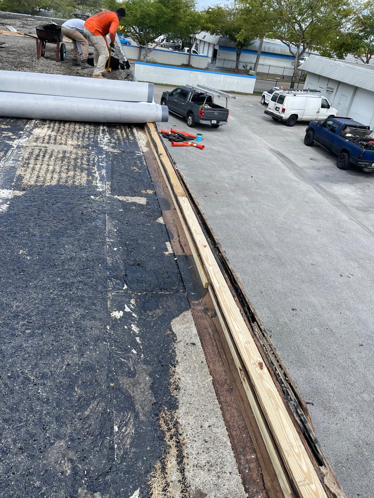 A worker repairs a flat roof near stacked supplies, with cars parked in a lot below.