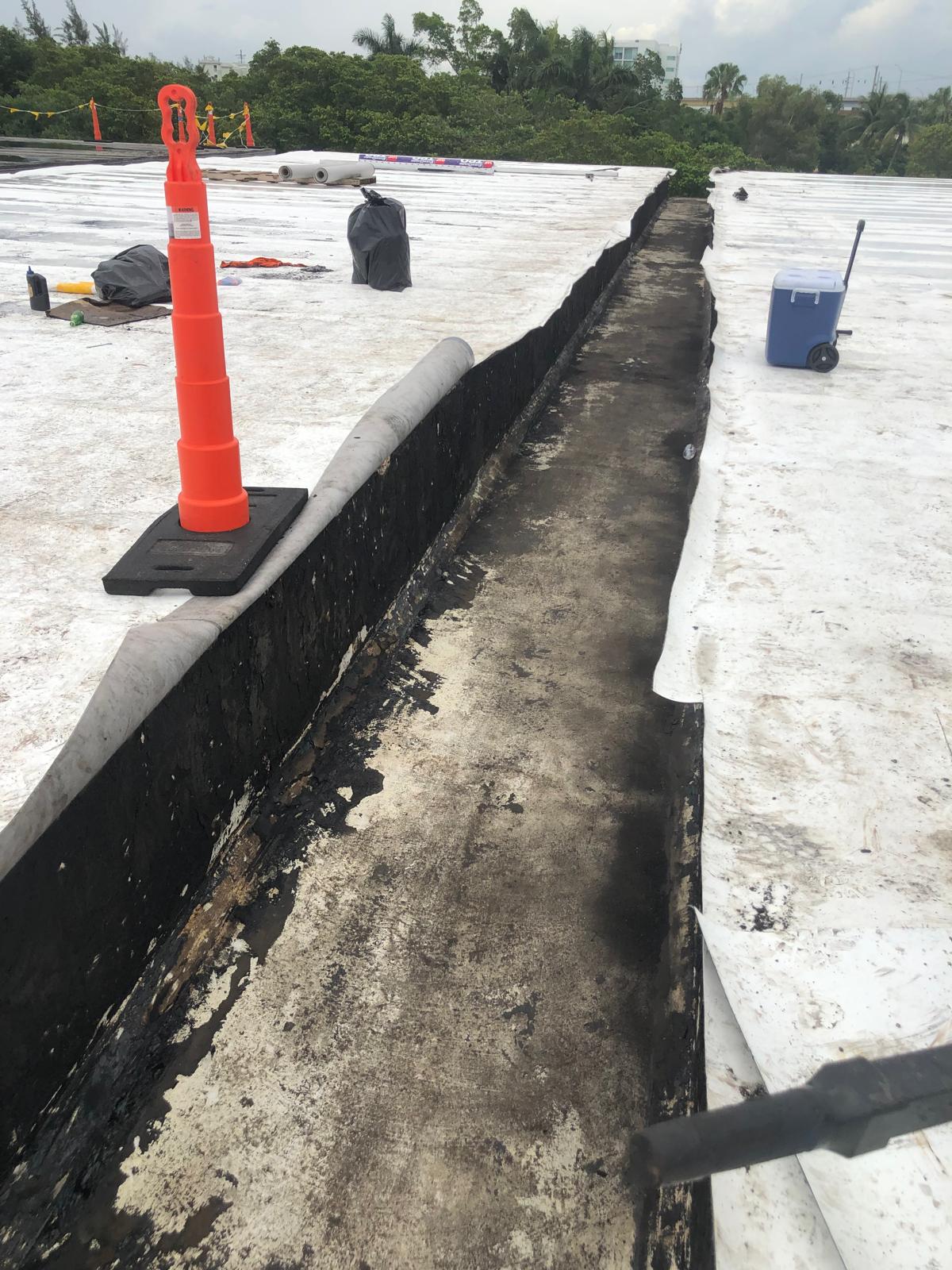 A construction site with a long trench in the roofing material and an orange safety cone nearby.