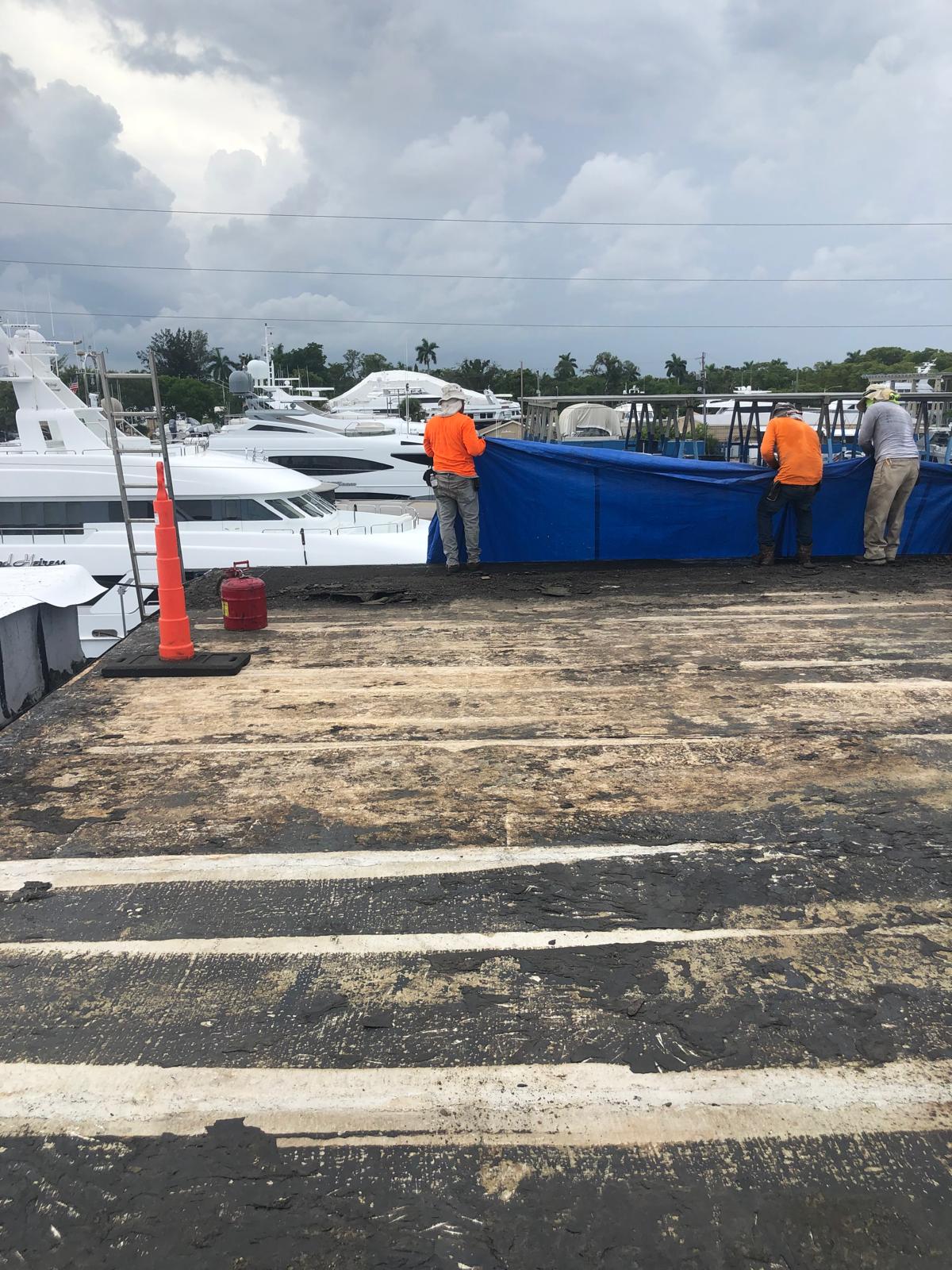 Three workers in orange vests stand by a blue tarp near yachts on a cloudy day, with traffic cones nearby.