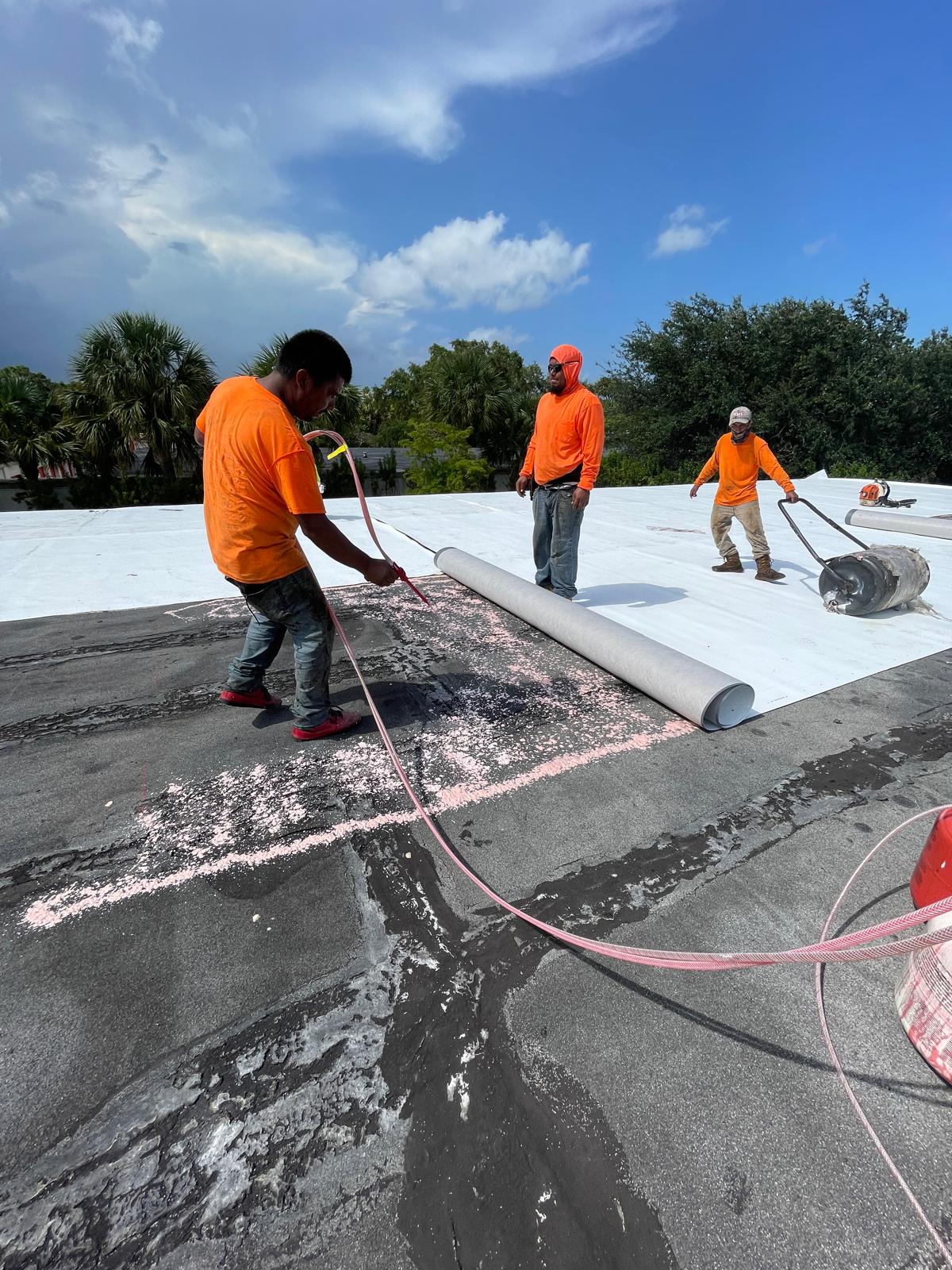 Three workers in orange shirts install roofing material on a flat roof under a partly cloudy sky.