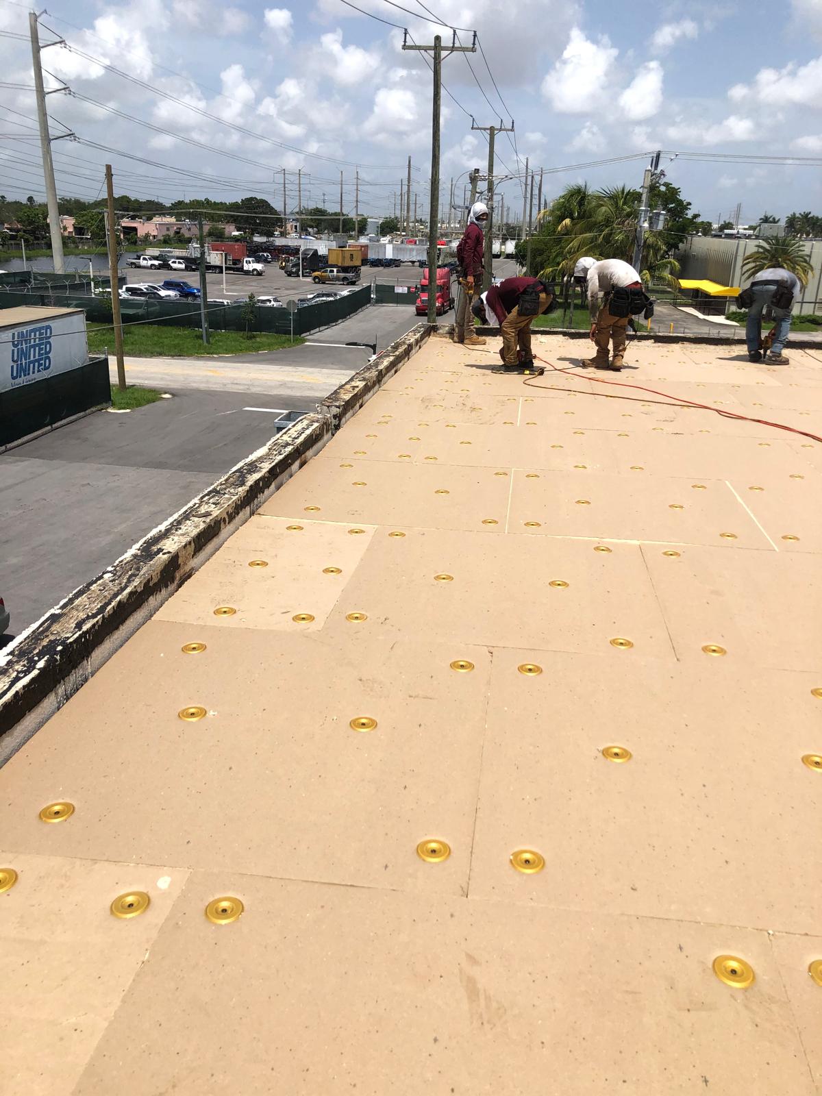 Workers installing roofing boards with yellow fasteners on a flat roof under a partly cloudy sky.