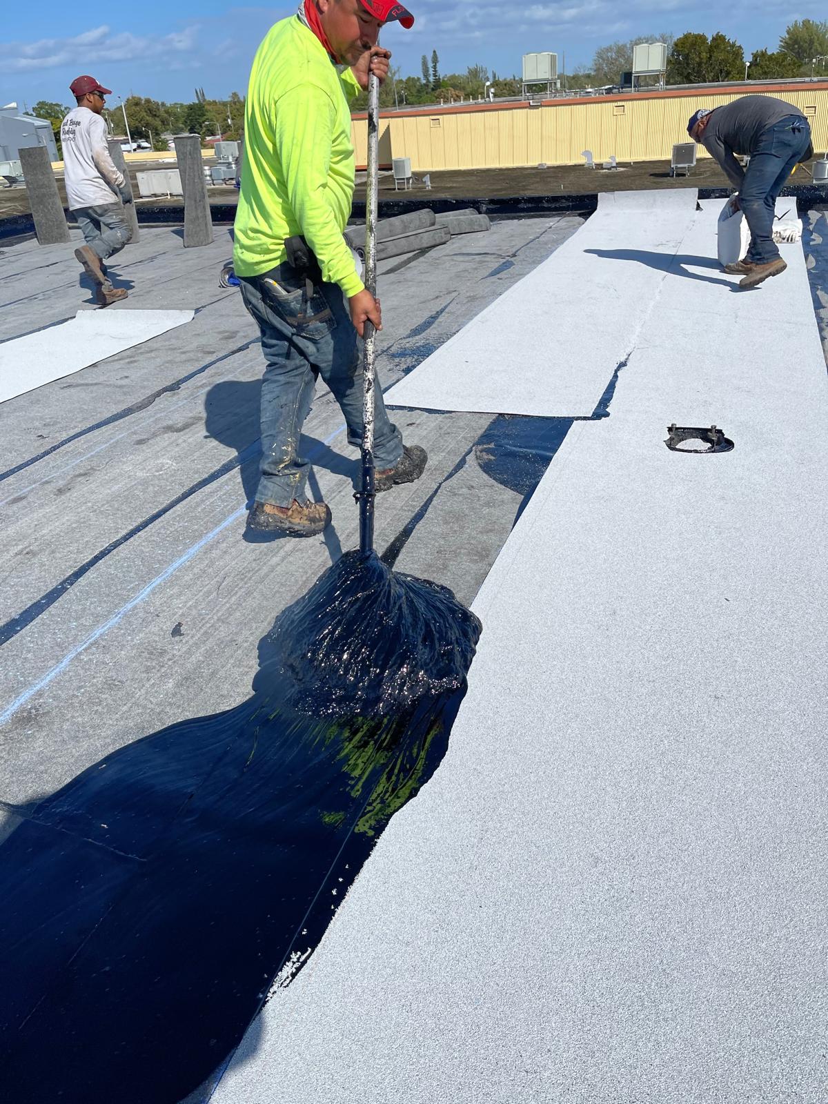 Worker in a neon shirt spreading black roofing tar on a flat rooftop with other workers nearby.