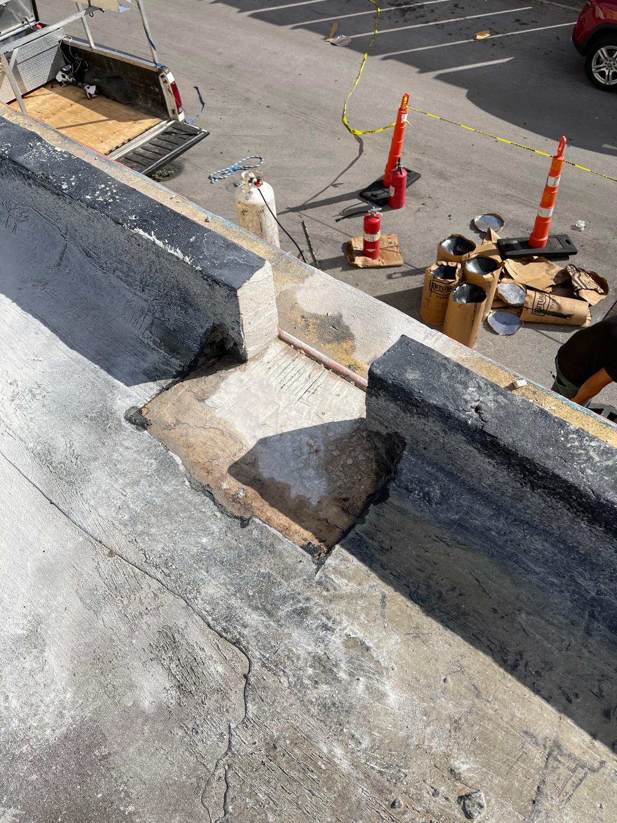 A damaged section of a rooftop with construction materials and orange cones below in a parking lot.