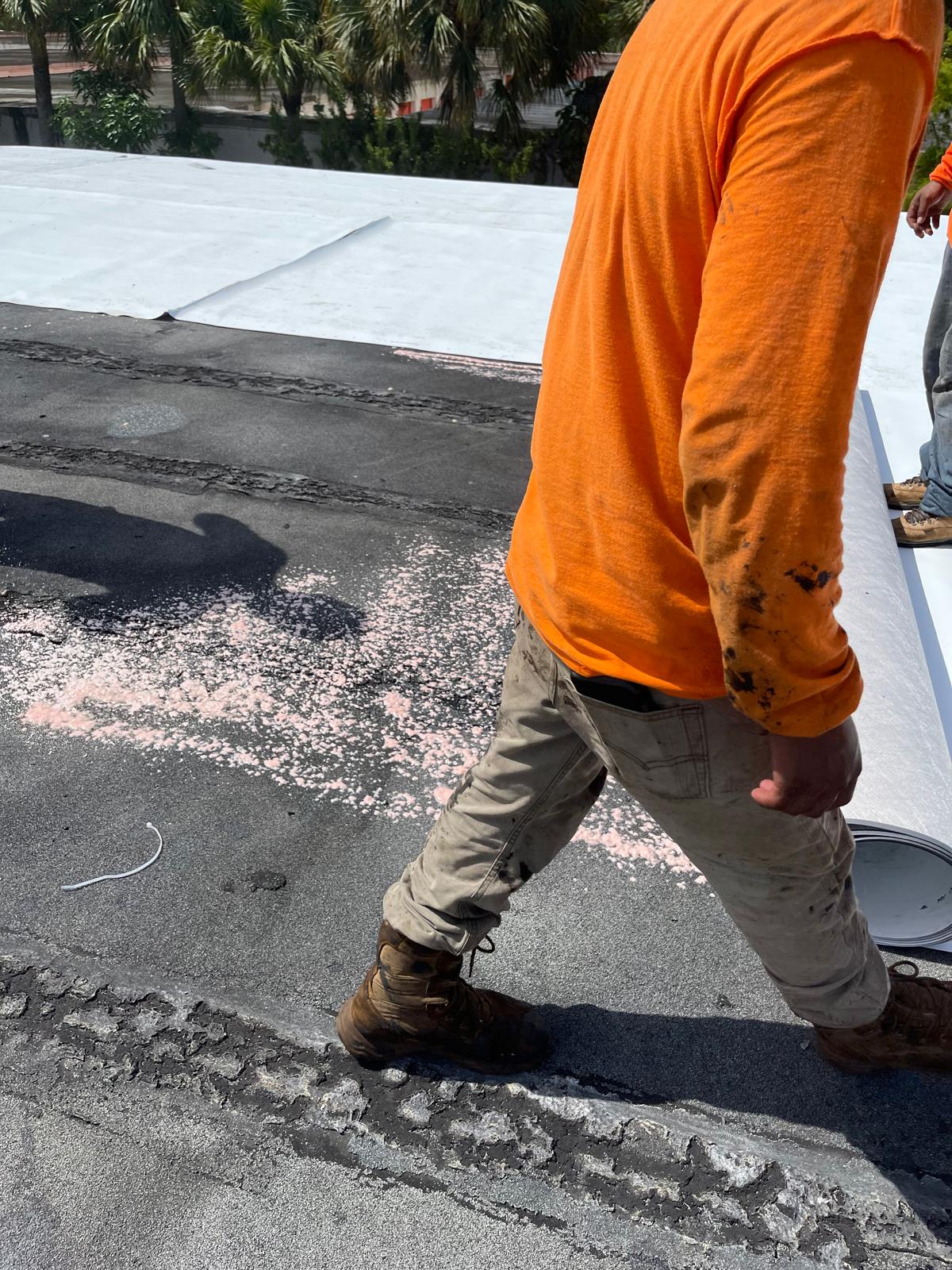 Worker in an orange shirt and tan pants walking on a rooftop with roofing materials and debris visible.